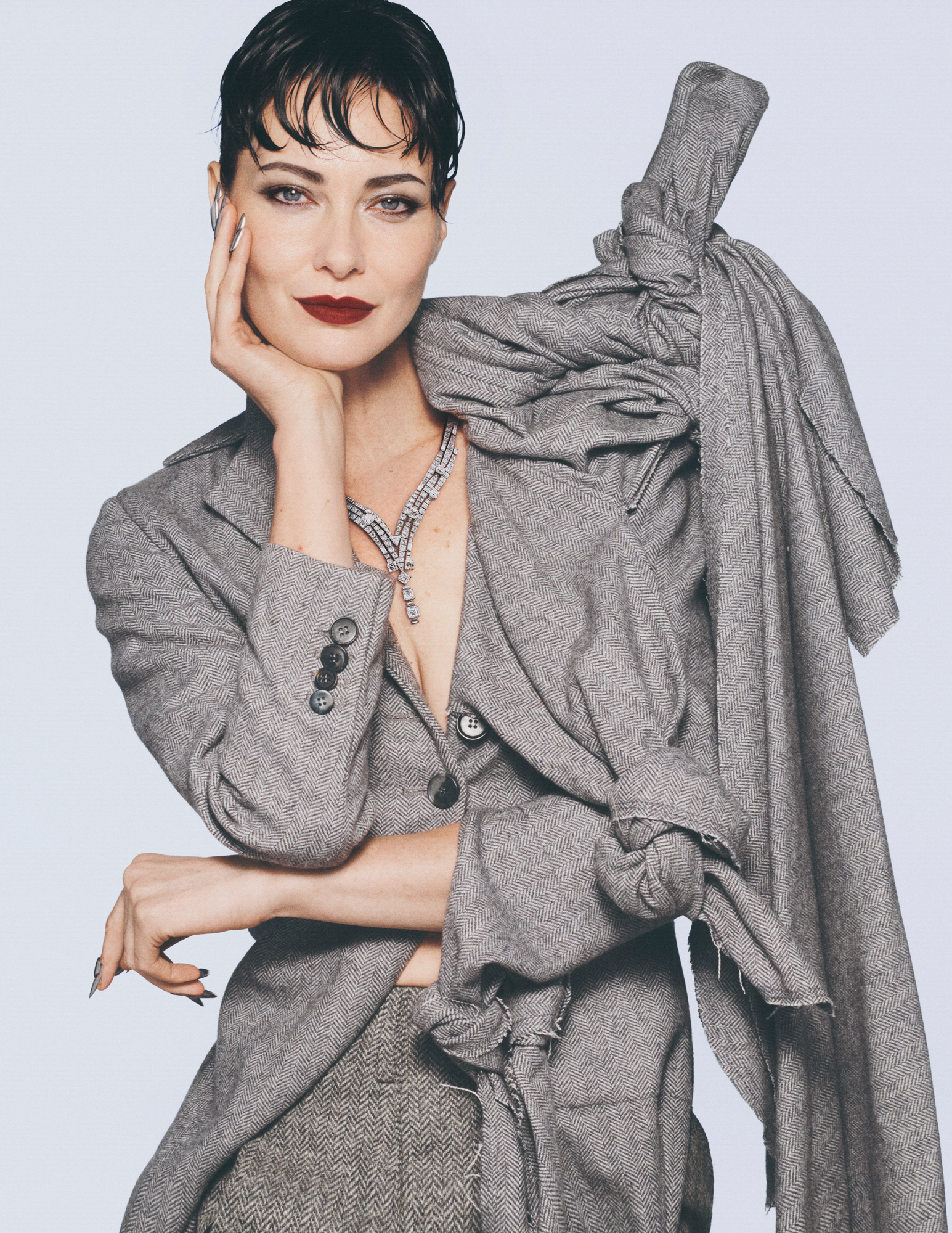 Shalom Harlow is wearing a grey suit jacket and grey slacks. She has a pile of grey fabric in knots on one of her shoulders. She is resting her chin on one of her hands. 