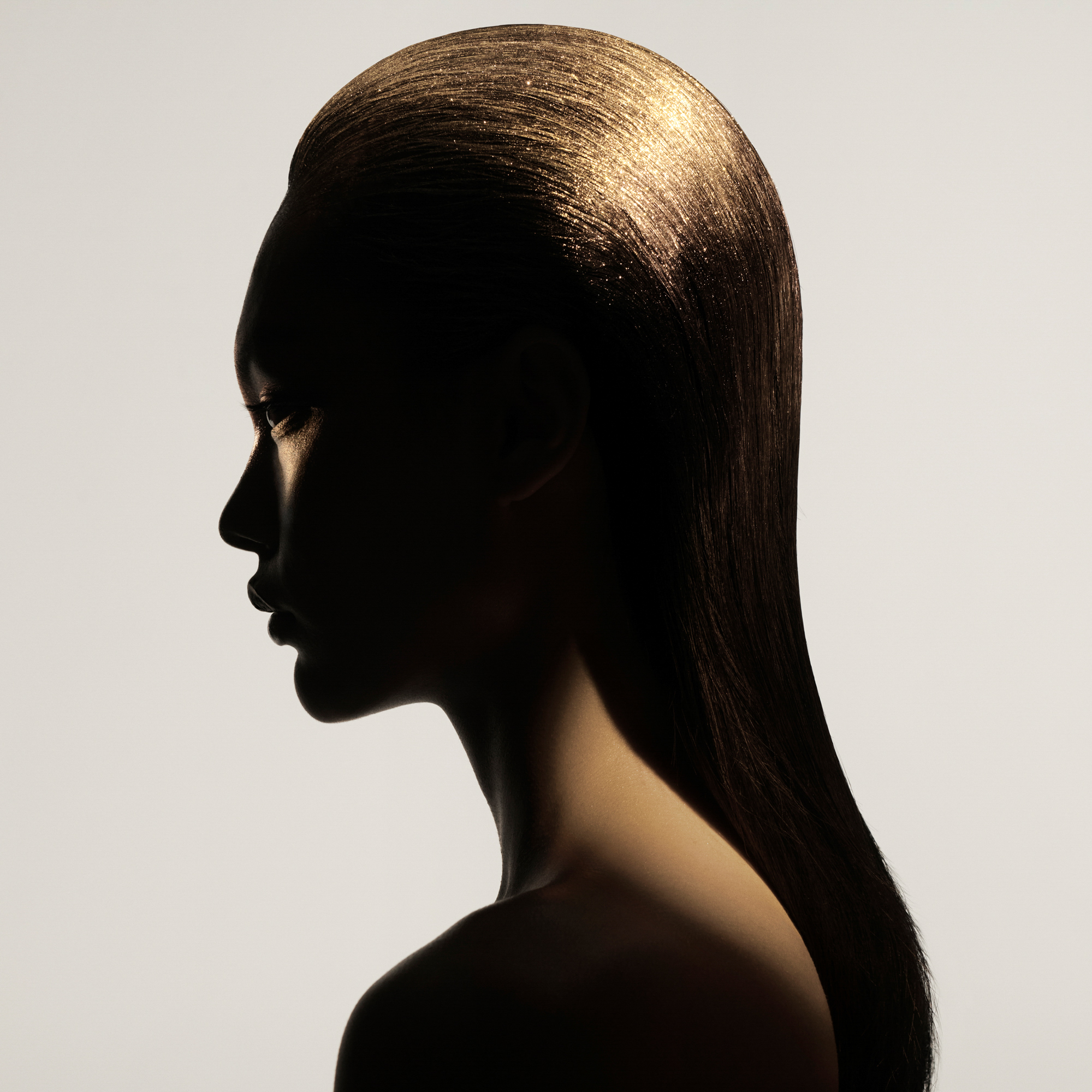A model is depicted in profile with her hair slicked back cleanly behind her back. The top of her hair is reflecting gold, and golden light is hitting her back and her eye. 