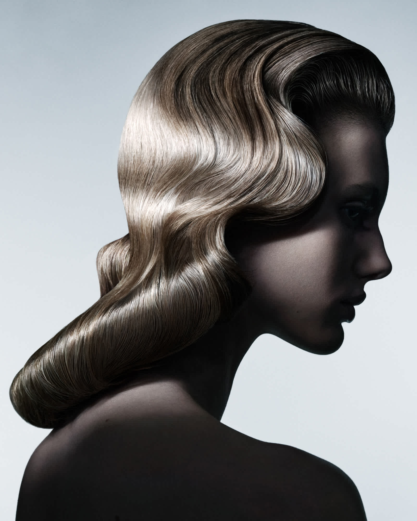 A woman with light blonde hair is seen in profile with a sculpted wavy look to the hair. Her face is hidden by shadow. The hair's shine is being illuminated from the left hand side of the frame.