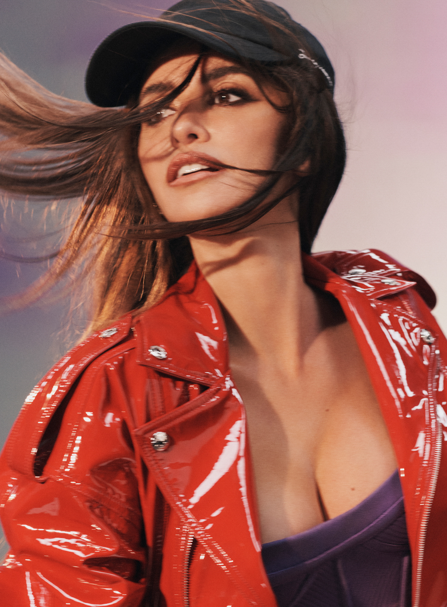 Penelope Cruz is looking off into the distance towards the left hand side of the frame with her hair blowing forward. She is wearing a shiny red leather jacket, purple bodice, and simple black baseball hat. 