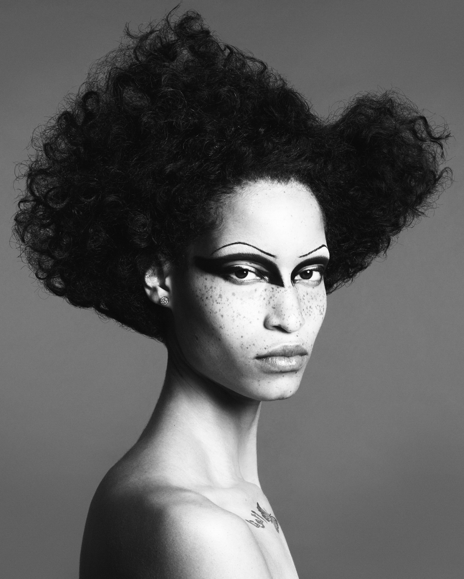A woman is depicted with her shoulders in profile, and her head slightly turned. She has thick angular black shadow in the eye sockets, and thin arched brows. The hair is in a loosely sculpted afro that undulates slightly on the top. 