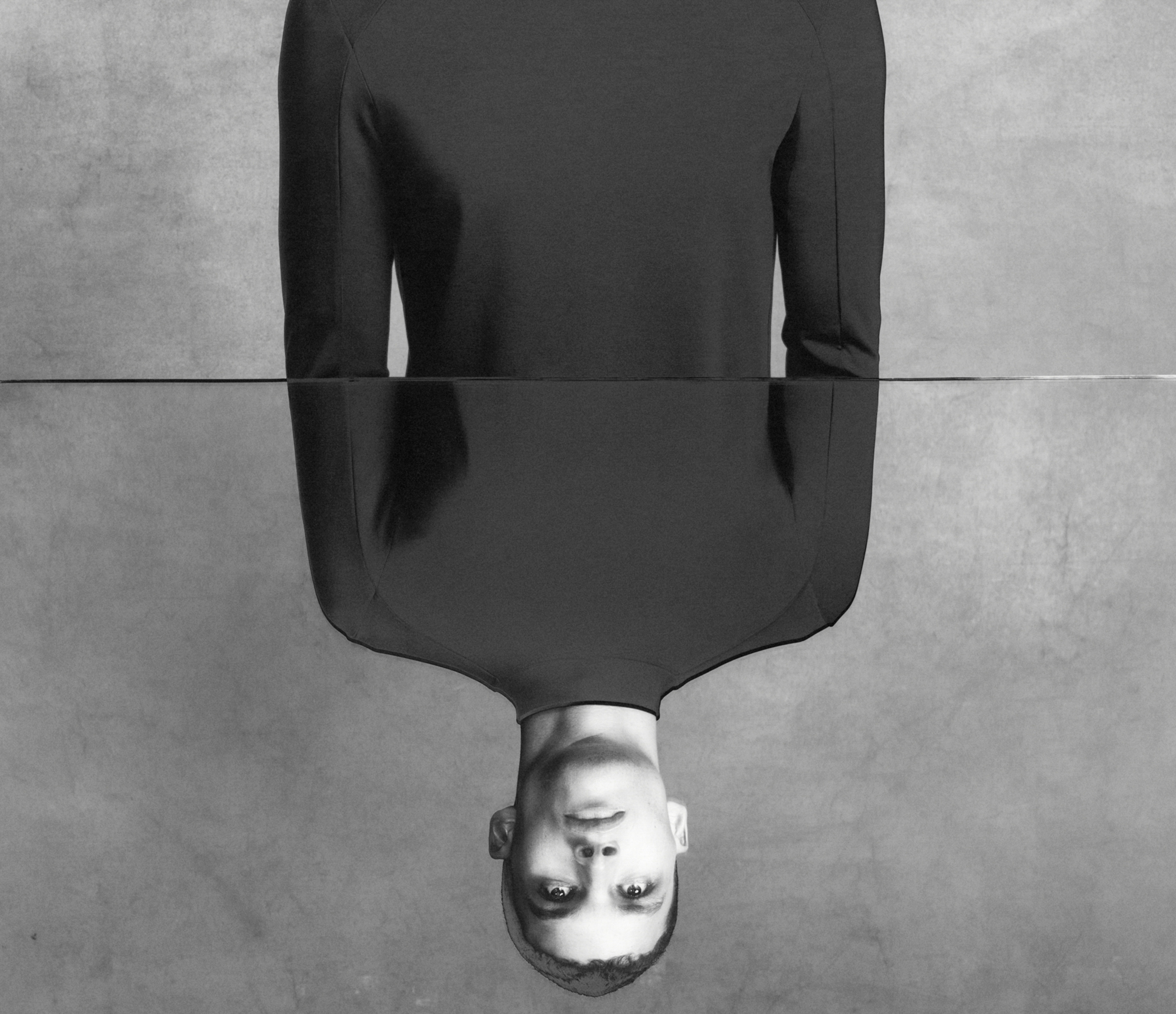 The frame of the image is split by a mirror surface. A man is standing in front of the mirror wearing a dark sweater, the mirror is cutting through the middle half of the man's torso. In the mirror's reflection you see the model looking up. The background is a textured grey. 