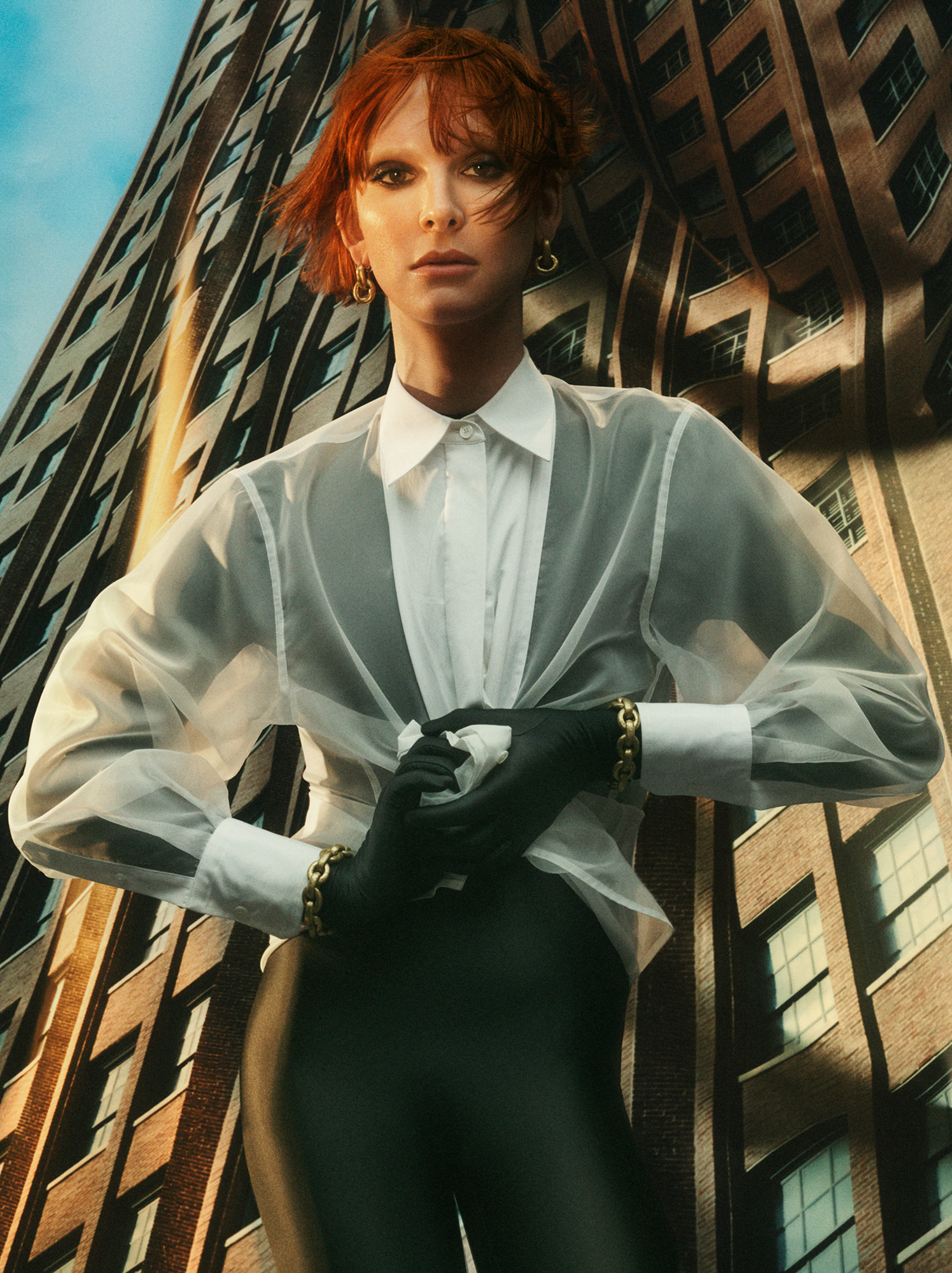 Hari Nef is centered in the frame, she is wearing a black body suit with a sheer white dress shirt over it. She is scrunching the fabric of the shirt into a ball near her stomach. She is wearing gold earrings and bracelets. The backdrop behind her is bent in certain places, and it is an image of a skyscraper with the perspective of being on the street and looking up. 