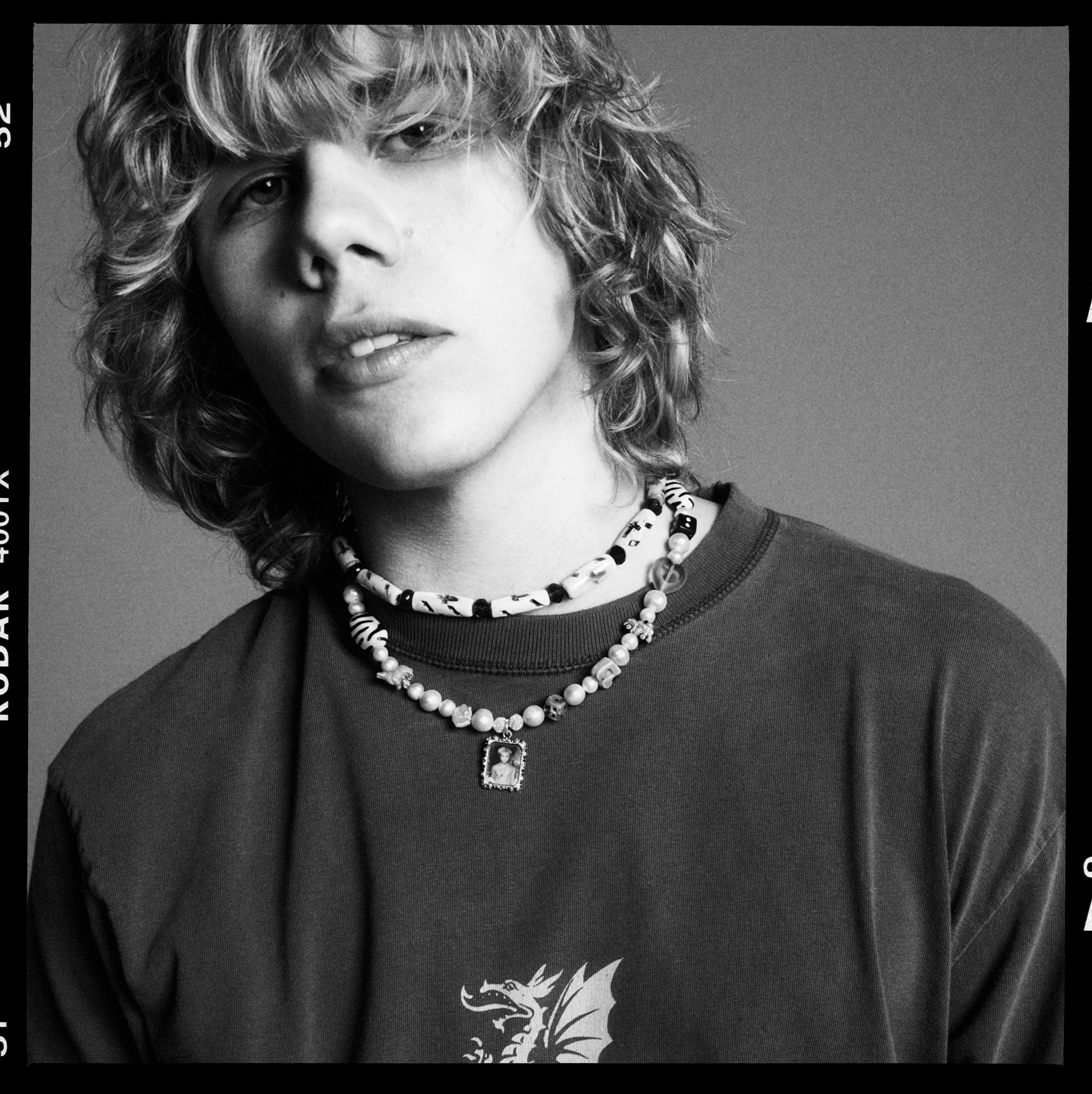 The image is a close up portrait of Kid Laroi. He is wearing a t-shirt with a dragon graphic on it, and has two beaded necklaces on. 