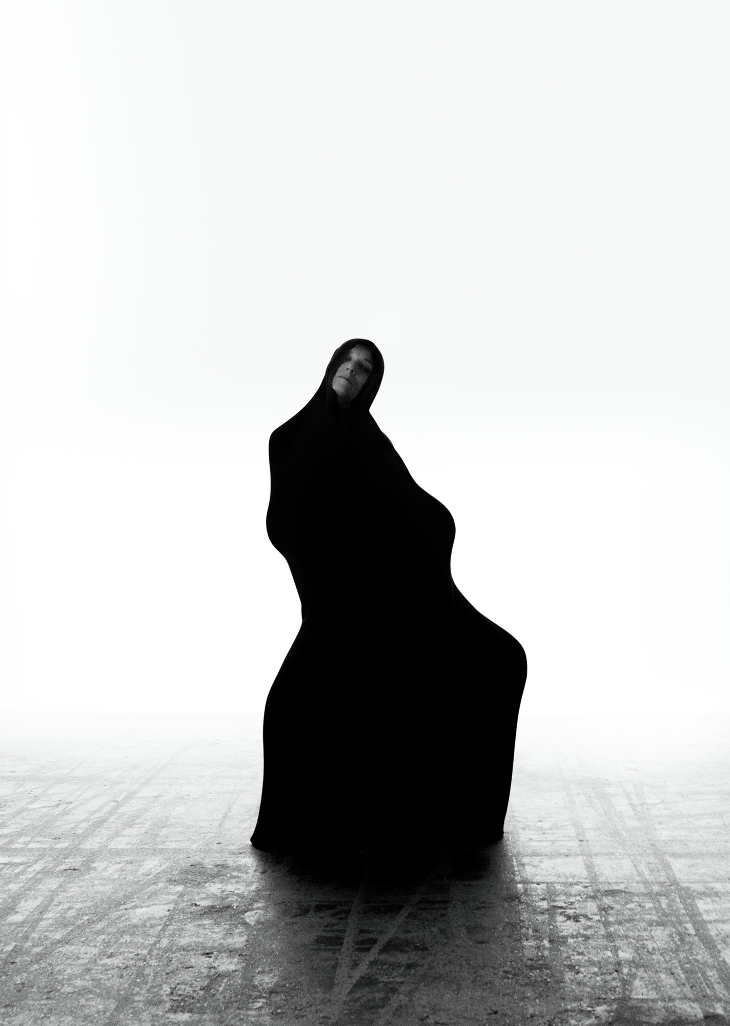 Figure wearing head to toe black is standing in the center of the frame. Light is flooding the background, and the floor is a grey concrete. 