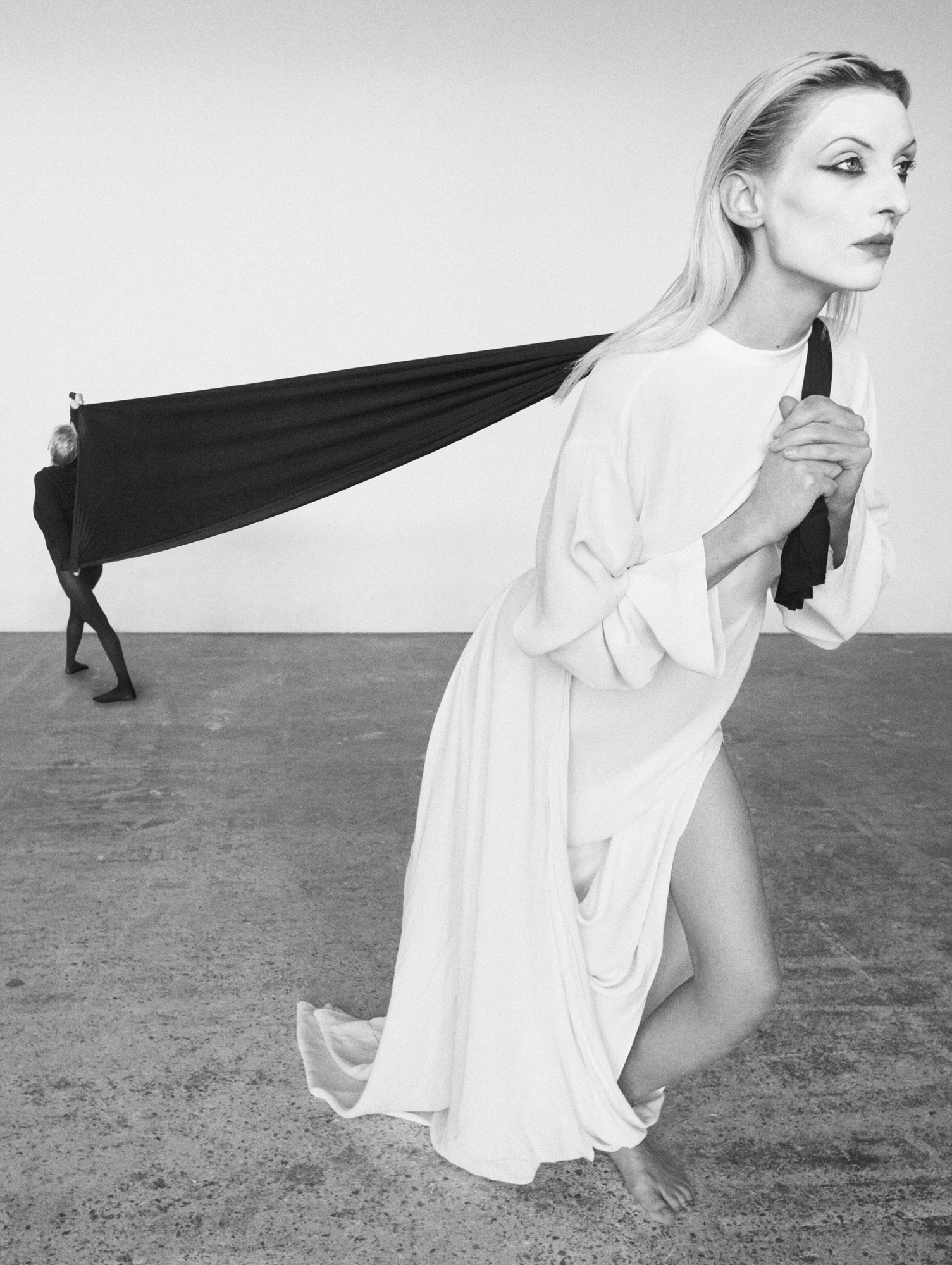 A woman is pulling a black rectangular fabric forward in the right hand side of the frame. There is a figured dressed in all black in the left hand side of the frame that is holding the fabric. A perspective shift is making the models size be skewed in the image. 