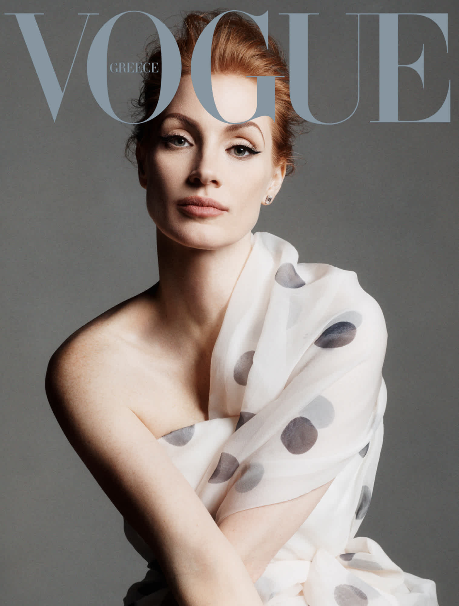 Cover of Vogue Greece with Jessica Chastain. She is wearing a white polka dot dress, and is looking directly at the camera with her arms crossed.