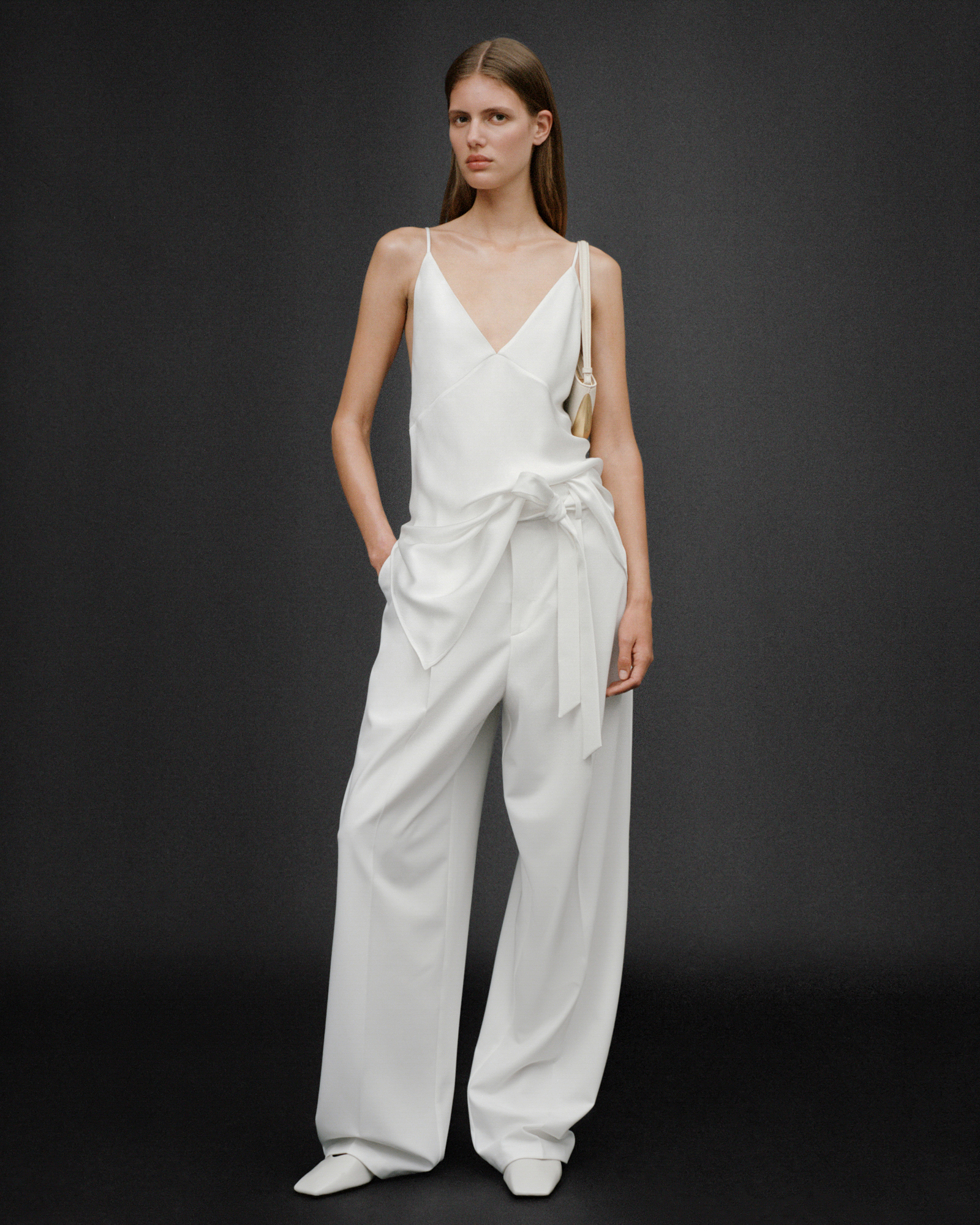 A woman is standing in an all white look against a black background. She is wearing a tank top, loose trousers, and a bag on the shoulder. 