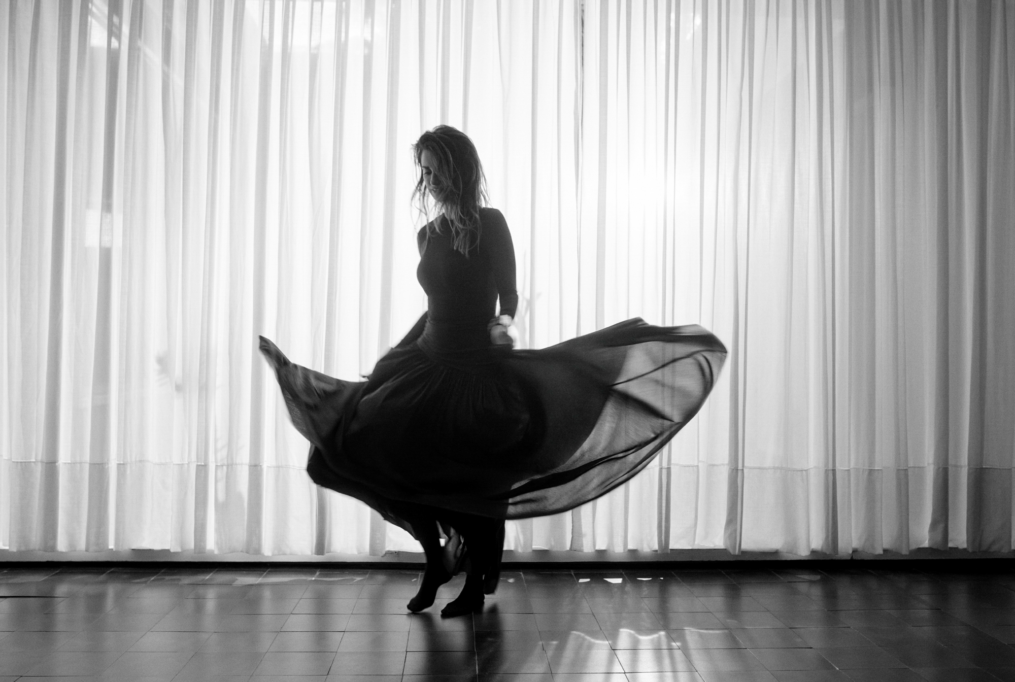 Penelope Cruz is twirling around in a full length dress. She is barefoot, and in a room with tiled floors and sheer curtains. 