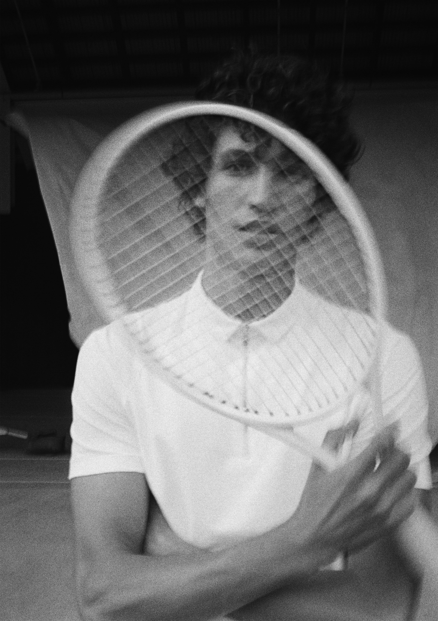 A man with dark curly hair is holding up a tennis racket in one hand and looking at the camera through it. He is wearing a white short sleeve shirt with a zipper. In the background there is a fabric backdrop blowing. 