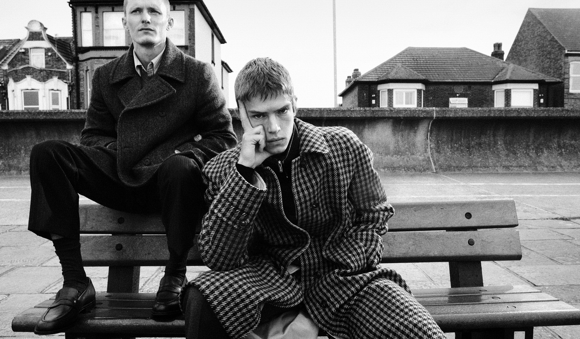 Two men are seated on a bench wearing coats and pants. One man is resting his head in one hand, while the man behind him is seated on top of the back of the bench and is looking in the distance past the camera. 