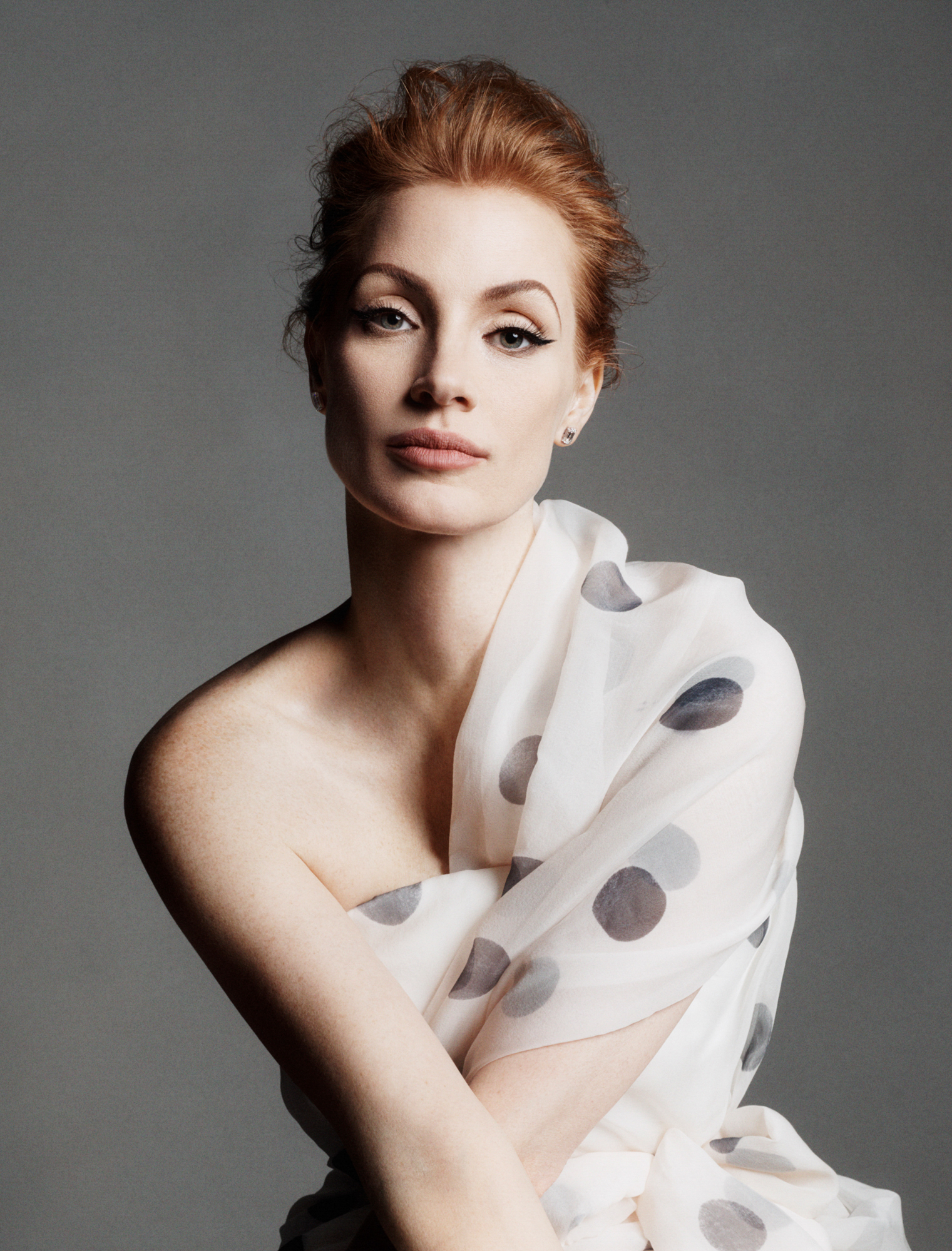 Portrait of Jessica Chastain looking directly at the camera with her arms crossed wearing a white polka dot dress. 