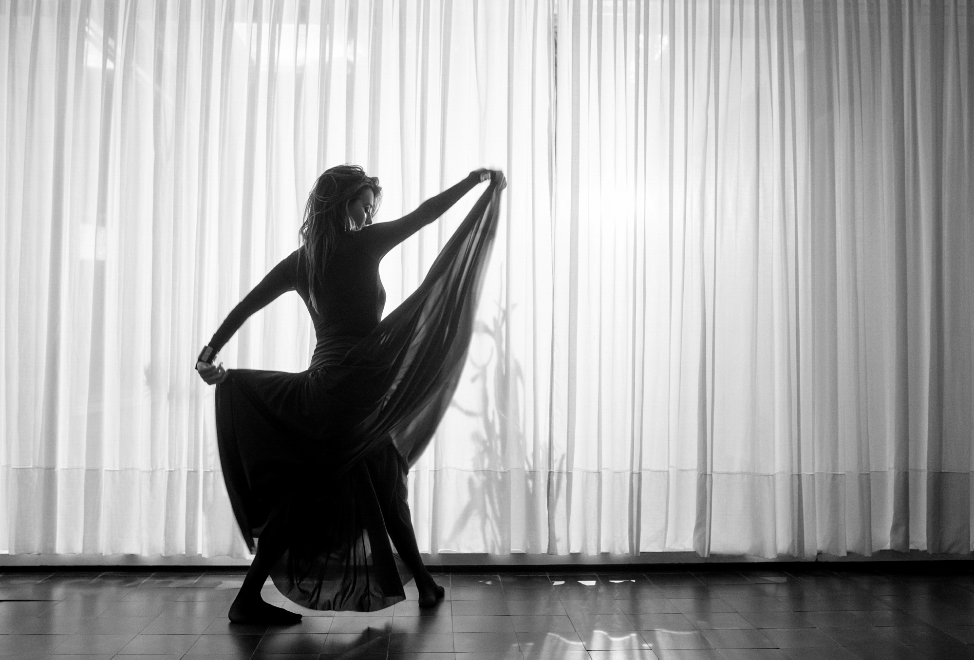 Penelope Cruz is holding sections of her full length dress up to allow for her feet to move freely. She is in an interior space with titled floors and sheer curtains in the background. 