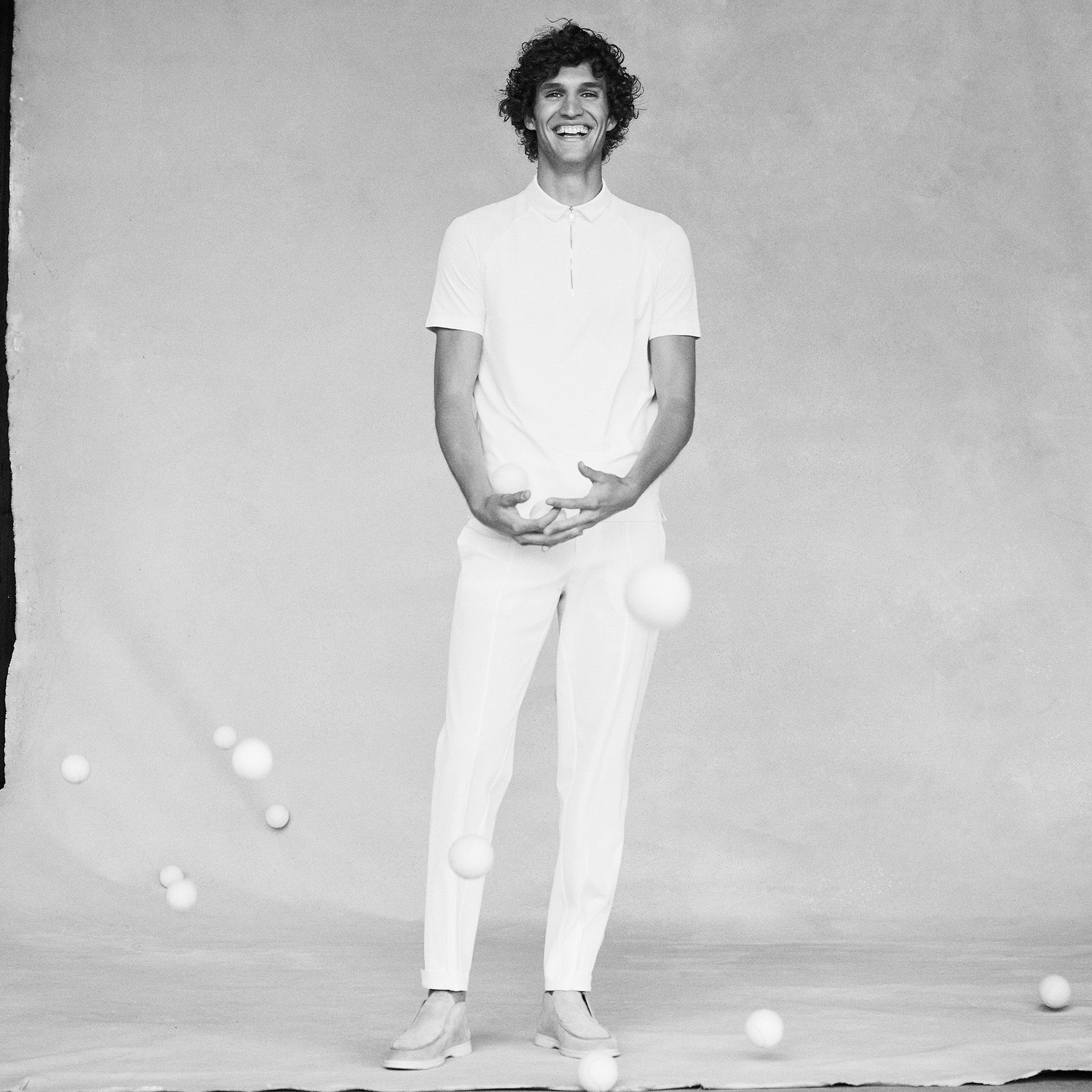 A man with curly dark hair is standing in the center of the frame. He is wearing a white shirt with a zipper, white pants, and light colored shoes. The backdrop is a light grey. The model is standing with his hands slightly together and smiling, all around him there are tennis balls bouncing around. 
