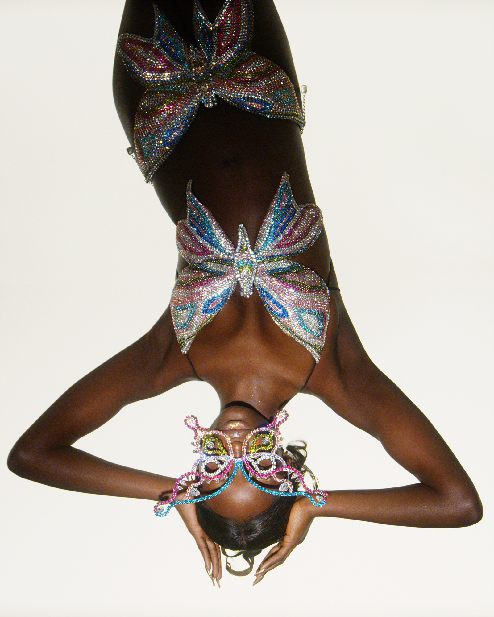A woman is depicted upside down in the frame, she has her hands on the sides of her head. She is wearing a halter top and skirt with jewels. Her face is partially covered by large glasses in the shape of a butterfly. 