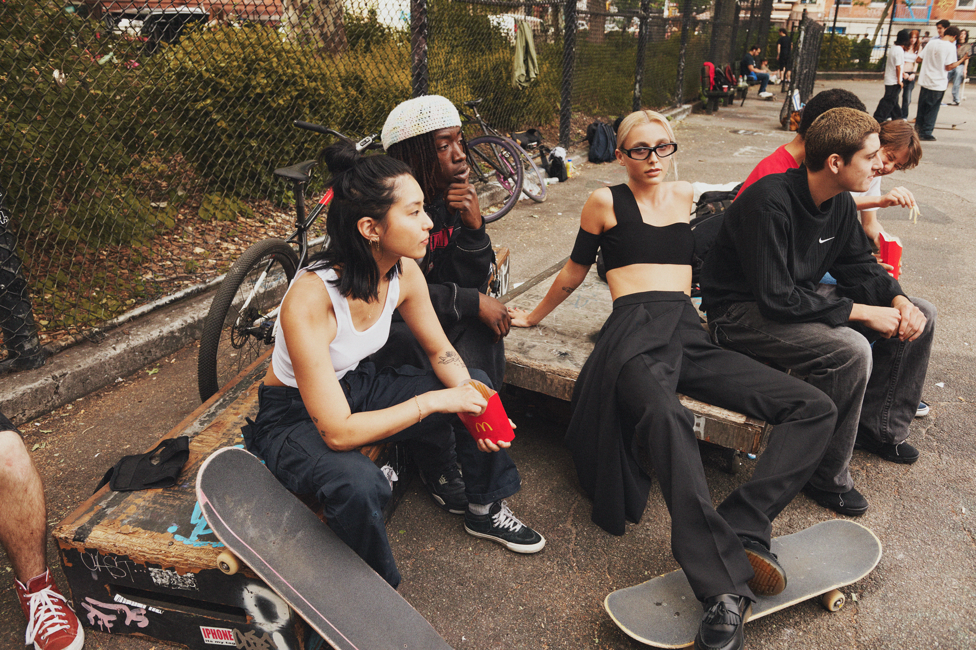 Emma Chamberlain is wearing a black halter top and black trousers. She is in a New York City basketball court, and with a group of young adults who have skateboards with them and are eating McDonalds French fries. 