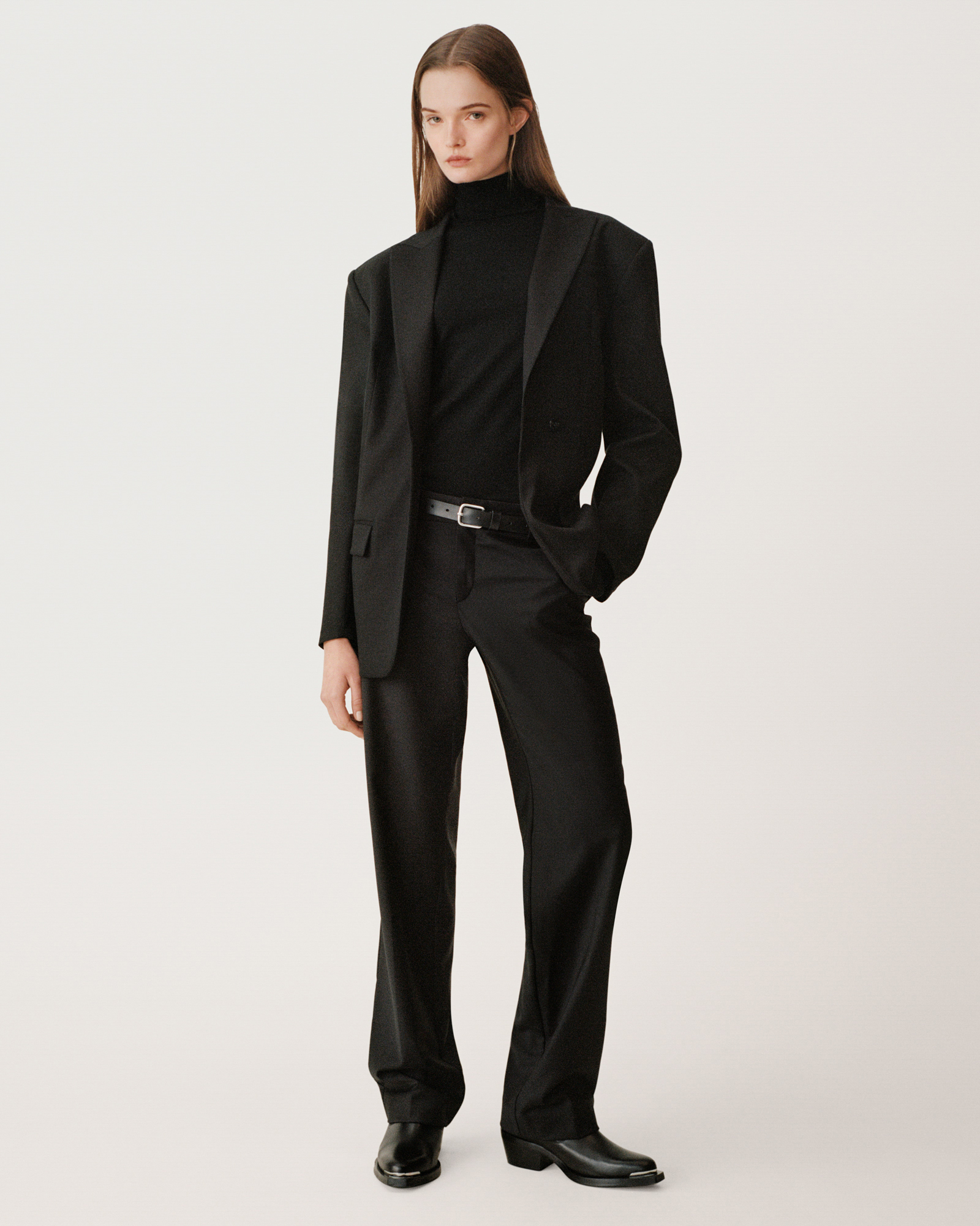 A woman is standing against a cream white background. She is wearing an all black look. A turtleneck underneath a blazer, trousers, and boots. She has one hand in her pocket while looking at the camera. 