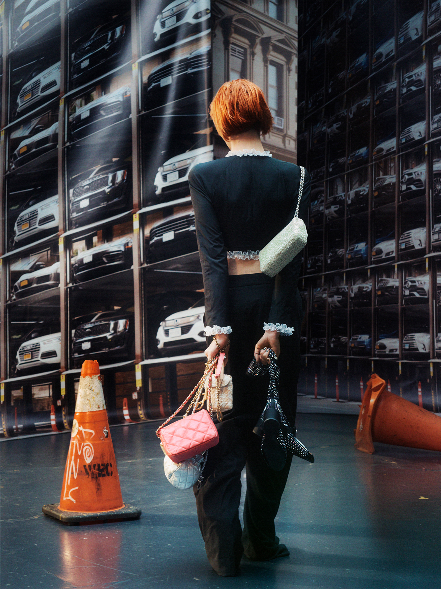 Hari Nef is standing in the center of the frame holding several quilted leather bags in one hand and a pair of black and silver shoes in the other. She is wearing a black crop top with white ruffles at the sleeves, neckline, and bottom of the garment. She is not facing the camera, there are orange caution cones on the floor, and in the background a backdrop of the elevated car parking structures where multiple vehicles are stacked on one another. 