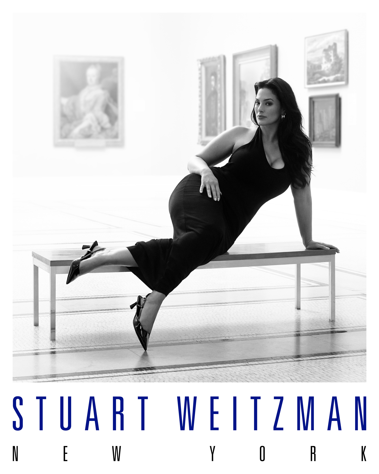 Ashley Graham is wearing a black dress with black pumps and is partially reclined on a gallery bench with paintings in the background. The Stuart Weitzman New York logo is featured below the image. 