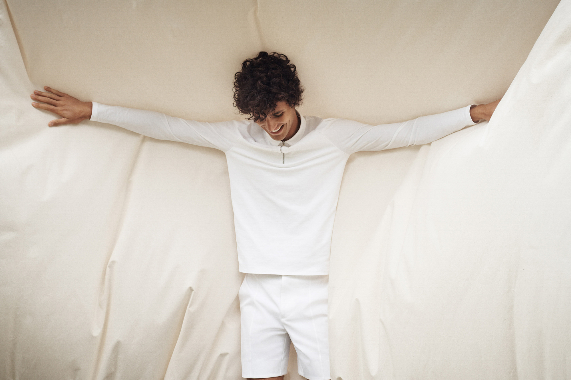 A man is standing within a cream colored fabric. His arms are outstretched, holding the cream colored fabrics on the sides. He is looking down and smiling, wearing a long sleeve shirt with zipper, and white shorts. 