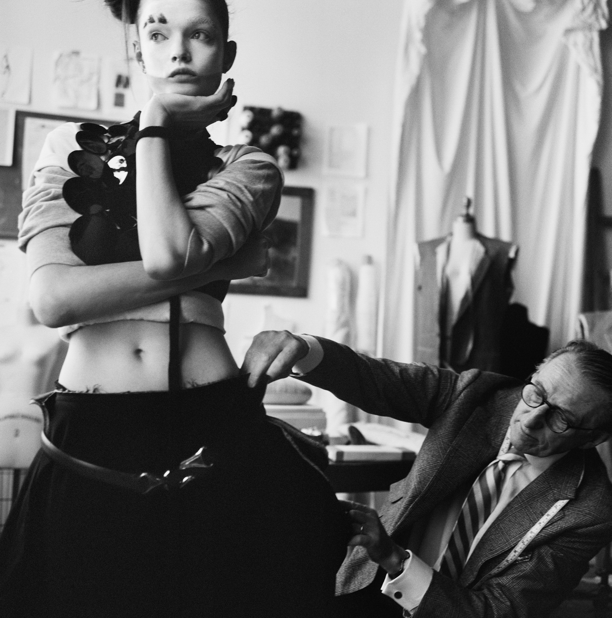 Lulu Tenney has her arms crossed and is holding on of her palms under her chin. A man to the right is holding up what looks to be a swatch and looking at it against that garment she has on. She is looking off to the left of the camera, and has mime looking makeup on. 