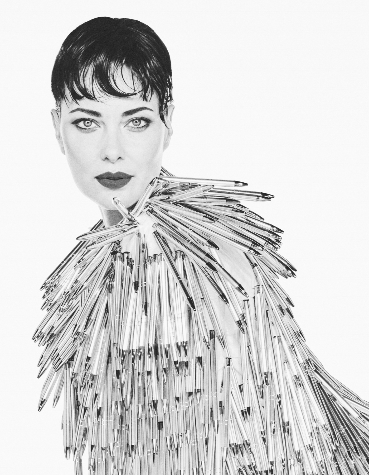 Shalom Harlow is leaning forward facing the camera directly. She is wearing a dress seemingly made out of pens. Her hair is short with a fringe. The background is a bright white. 