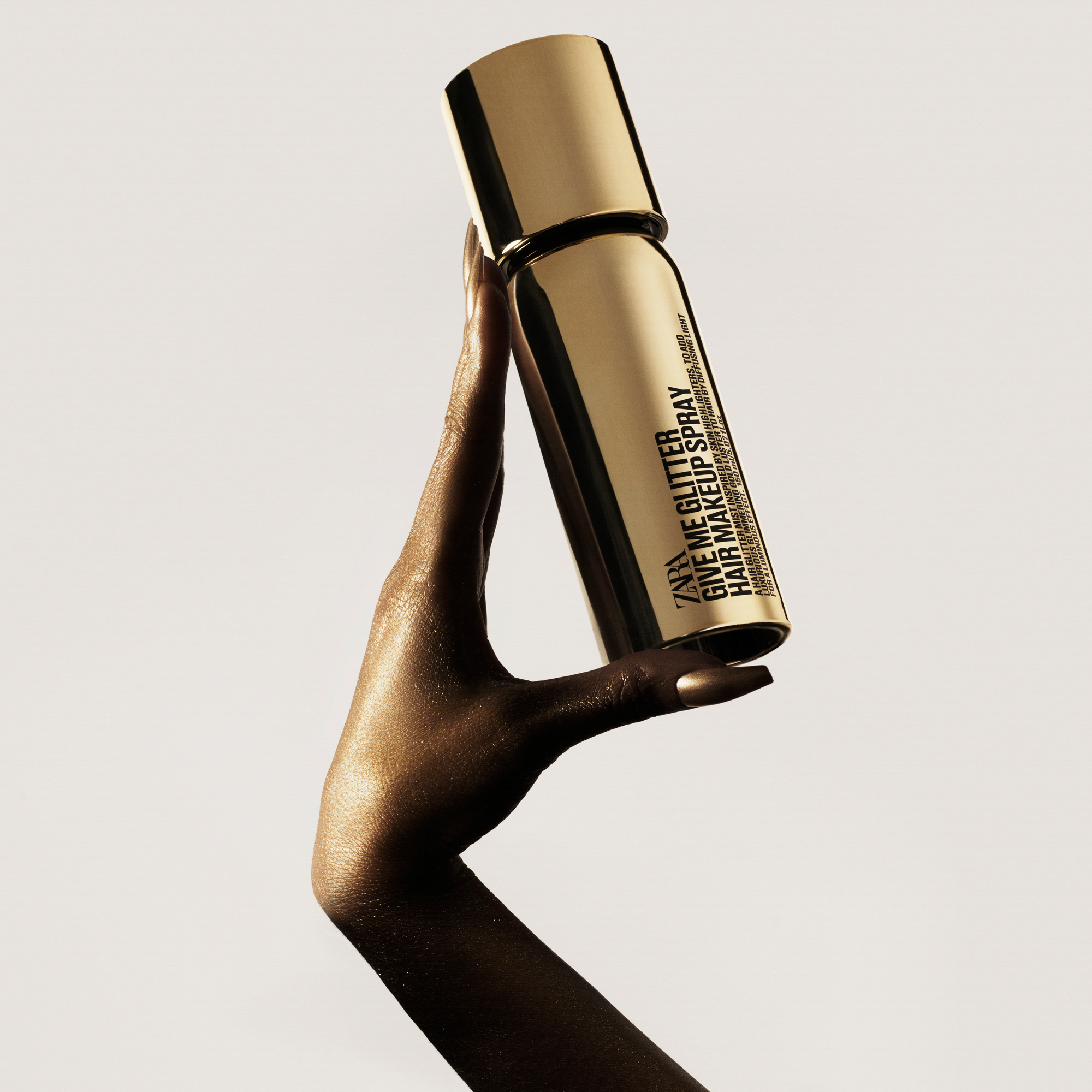 A model's hand that is covered in gold paint with gold nails is holding up a Zara hair makeup spray. The hand and bottle is shot against a white background. 