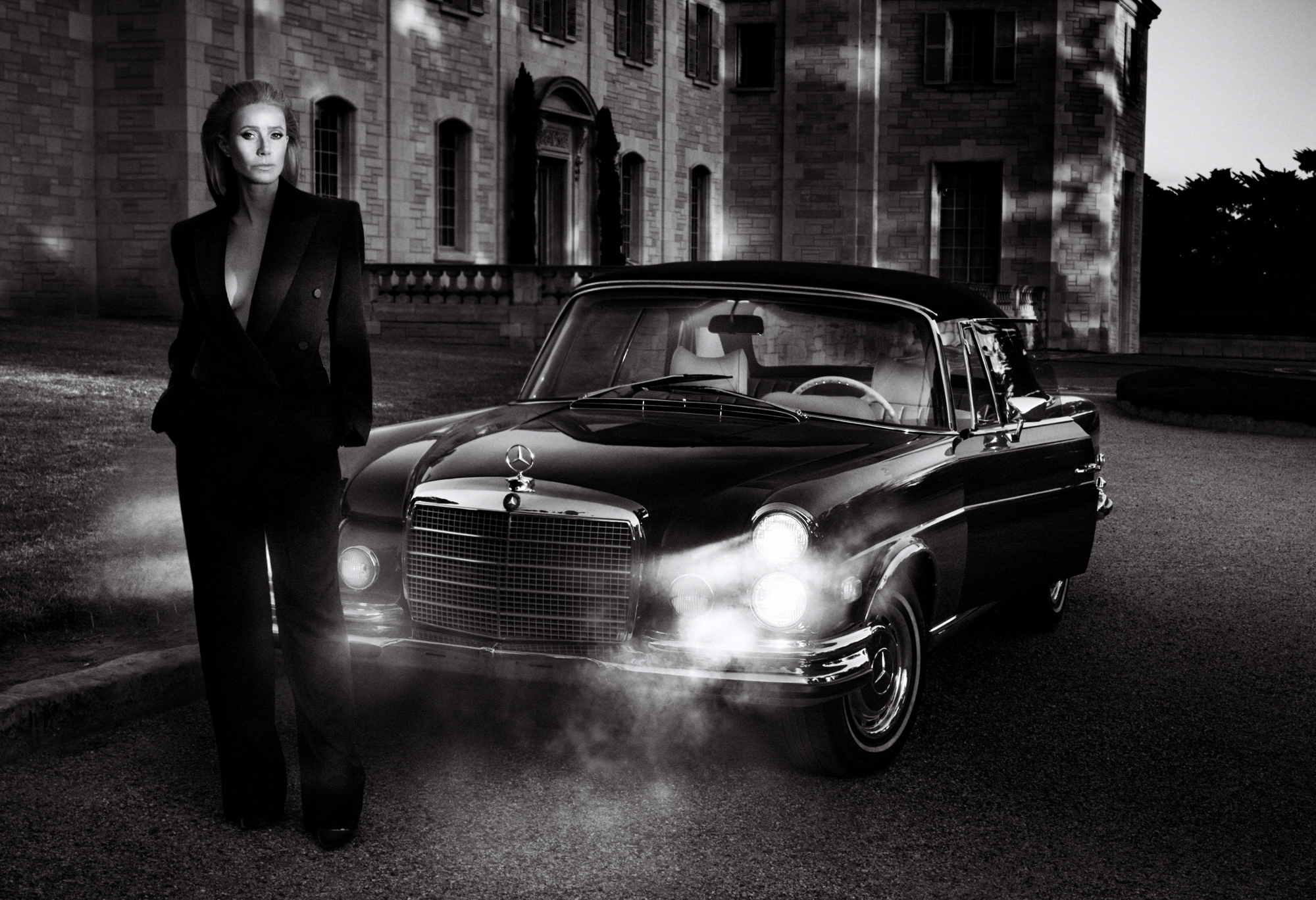 Gwyneth Paltrow is standing in an estate driveway wearing an all black suit. She is standing in front of a vintage Mercedes Benz car with its lights on illuminating mist. 