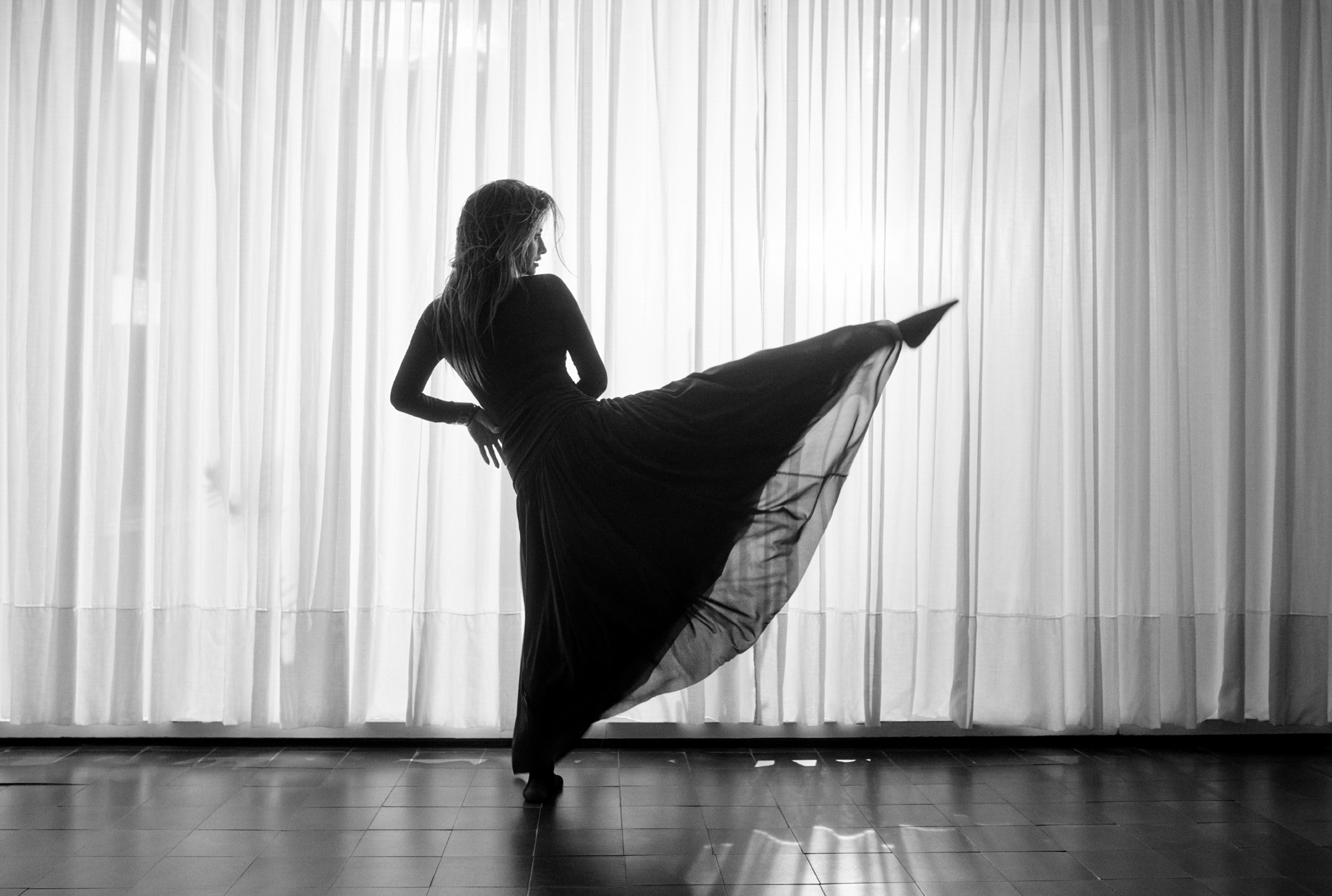 Penelope Cruz is kicking up one of her legs forward while wearing a full length dress. She is facing the right side of the frame. She is within a room with tiled floors and sheer curtains in the background. 