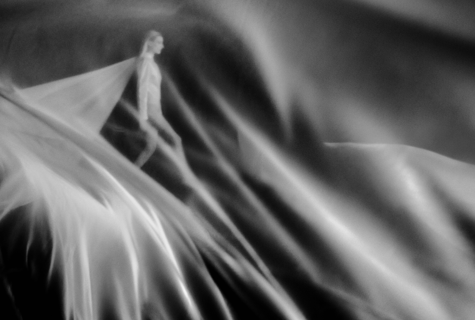 A figure with fabric attached to her back is walking forward. The entire frame is filled with flowing sheer fabric. 