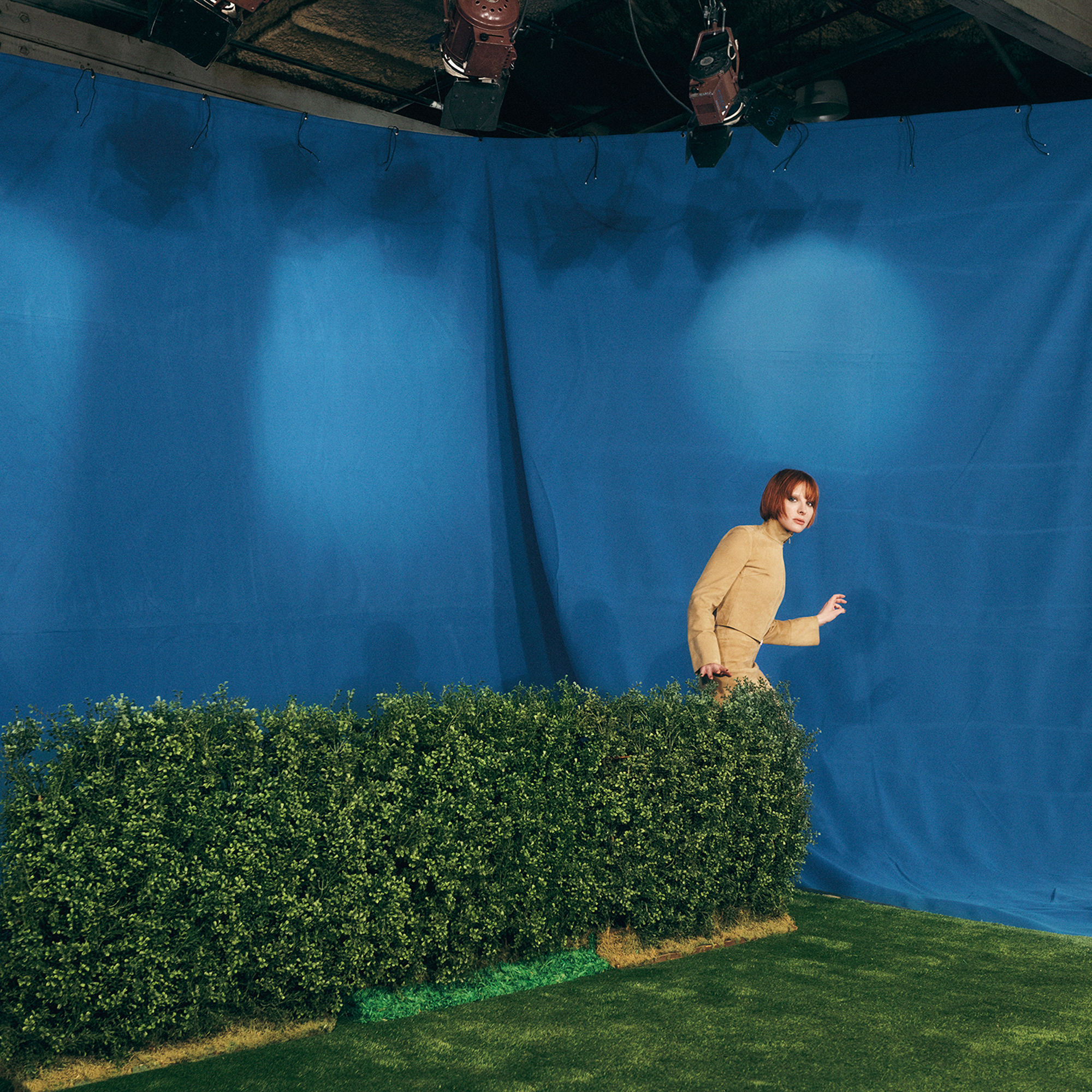 Hari Nef is wearing all beige with an orange bob. She is visible at the edge of a green bush, mid step as if she is sneaking around. The floor is covered with green astro turf, and a blue backdrop that is illuminated with spotlights. 