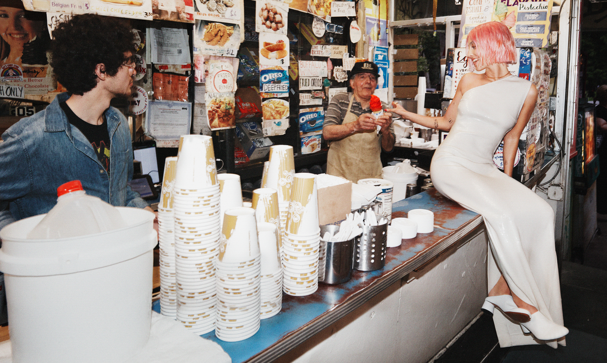 Emma Chamberlain is wearing an off the shoulder silver dress, she is sitting on the counter of an ice cream shop stand. She is turning around to grab a red colored ice cream cone. 