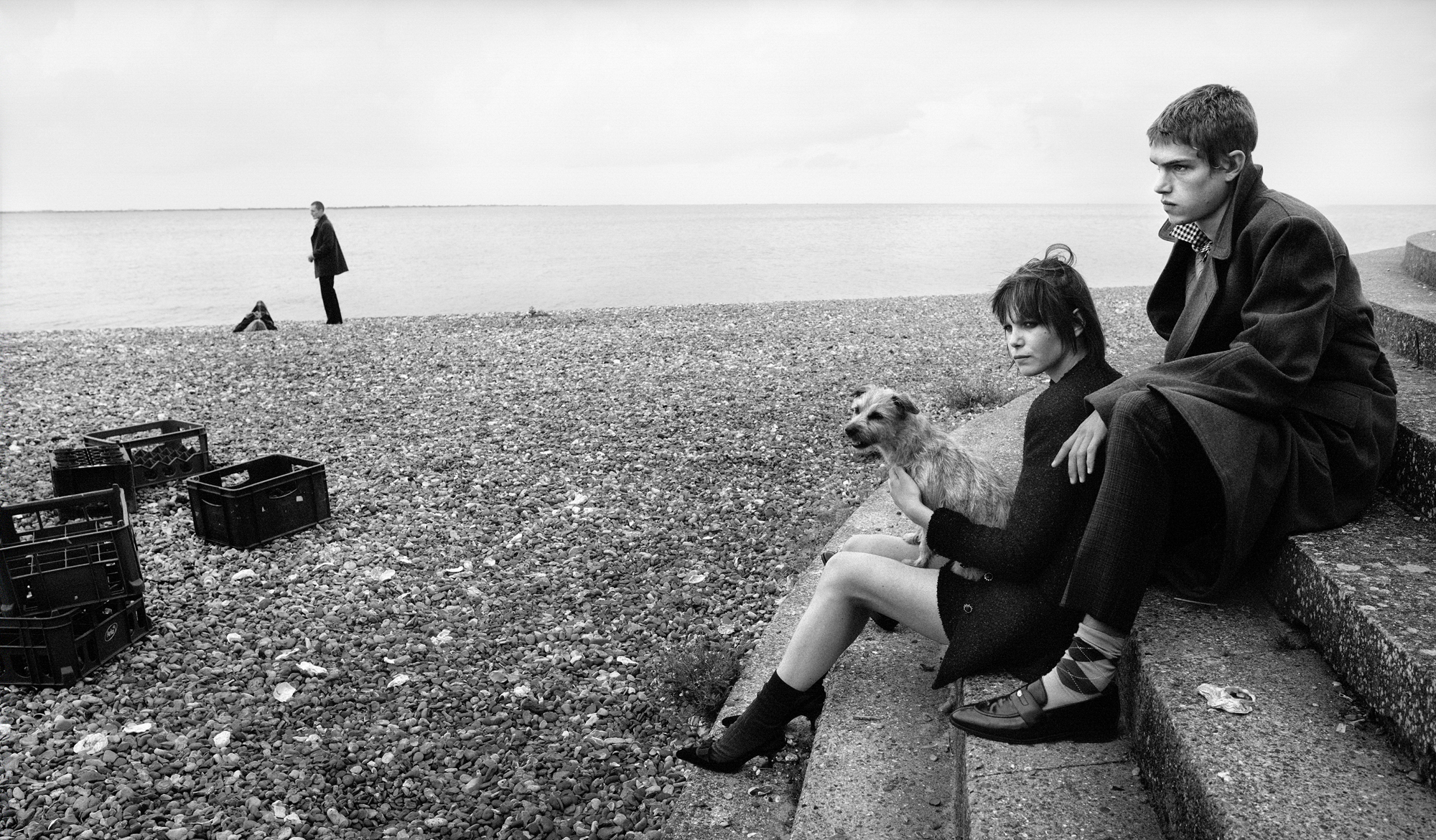 A young man and woman are seated on a stone staircase that looks out onto the sea. A small dog is with them, in the distance you can see milk crates on the rocky beach and a man and child visible near the water. 
