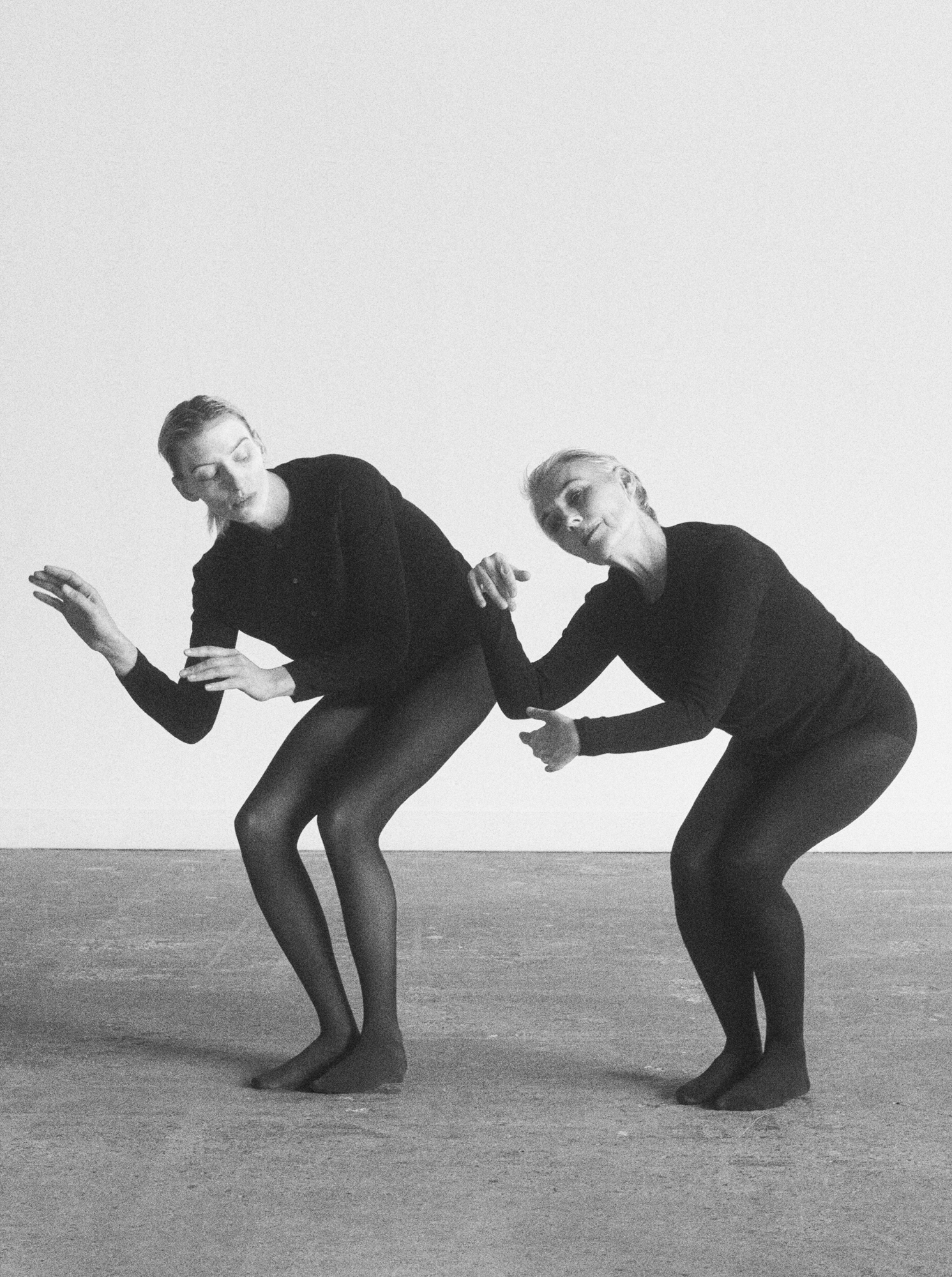 Two models are squatting with the torsos leaning forward. Both have their forearms angled up towards the ceiling. They are both wearing fitted black garments. 