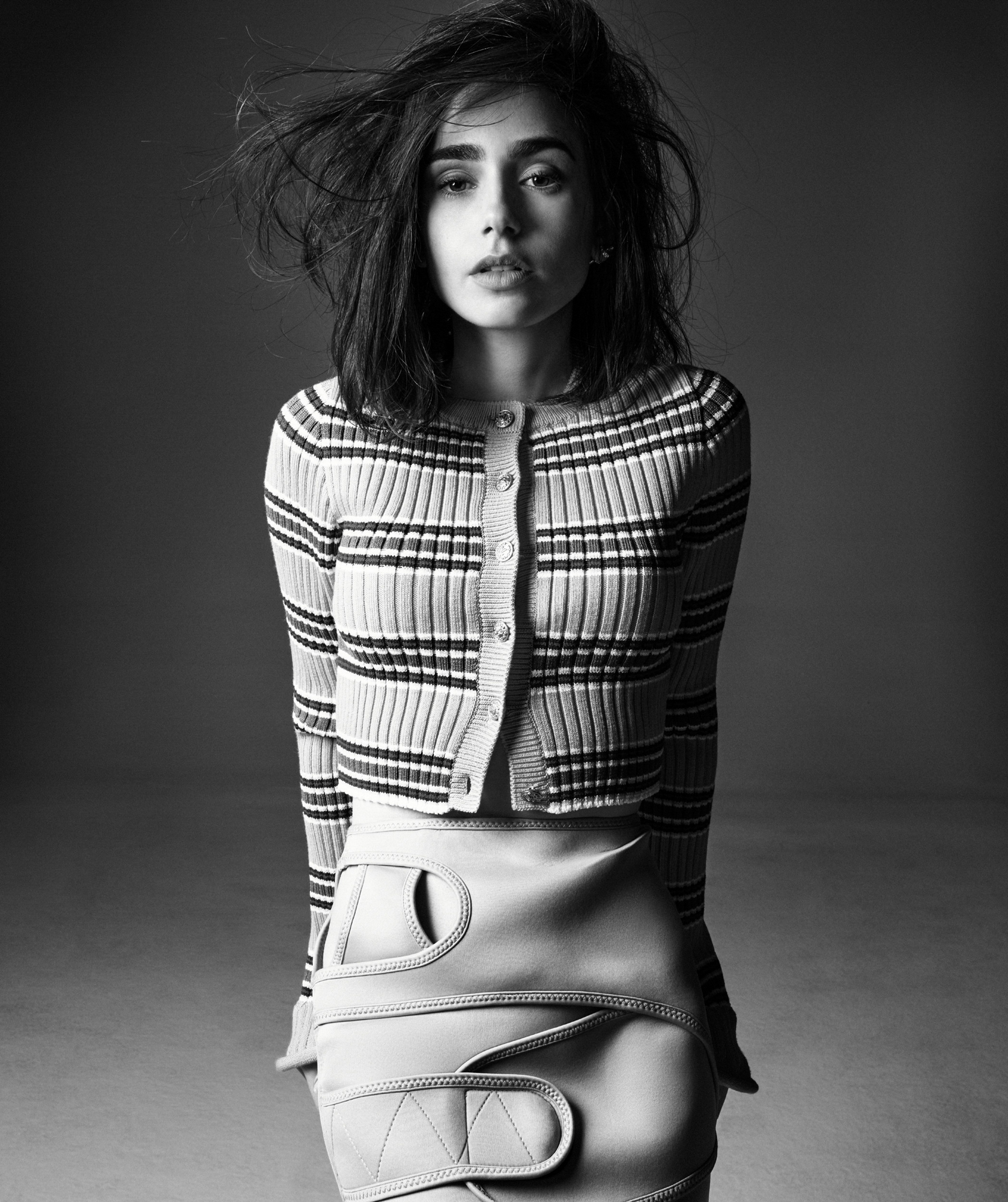 Lily Collins is standing in front of a dark grey studio background. She is wearing a striped knit sweater. Her skirt is a neoprene looking garment with straps. 