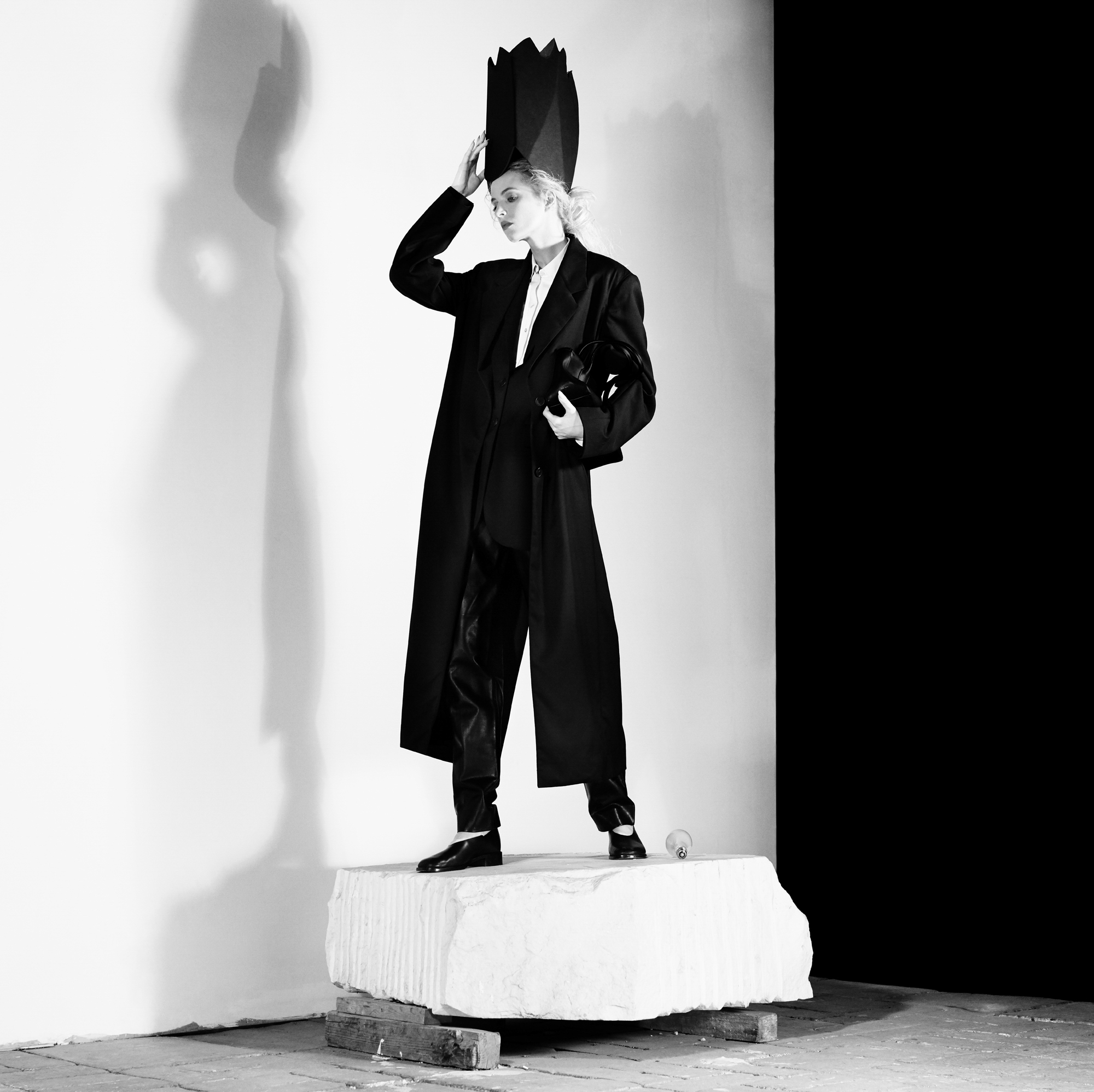 A model is standing atop a stone slab. She is wearing a black trench coat, white dress shirt, black pants, and leather shoes. She is holding a large crown like hat atop her head. Her eyes are looking down. 