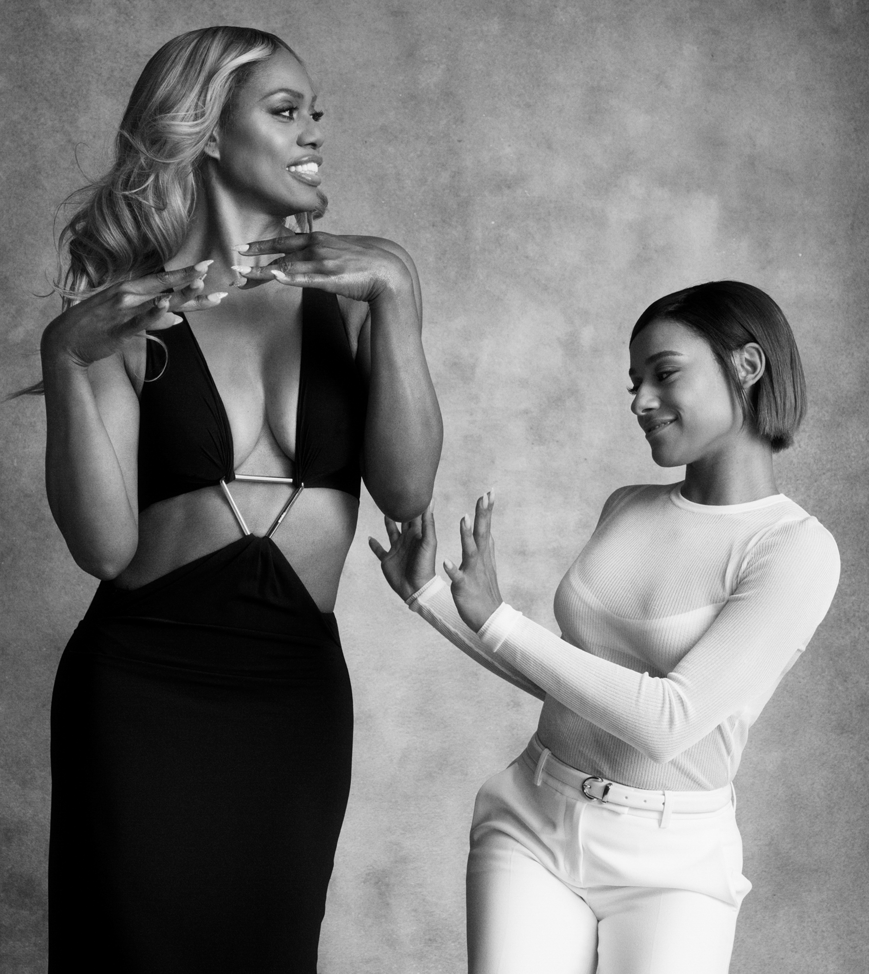 Laverne Cox and Ariana DeBose are standing next to each other on a textured grey background. Laverne is wearing a black dress with cut outs on the sides of the torso, she has her hands raised below her chin with the back of the hands facing up. Ariana is wearing all white and has her hands gesturing up towards Laverne. 