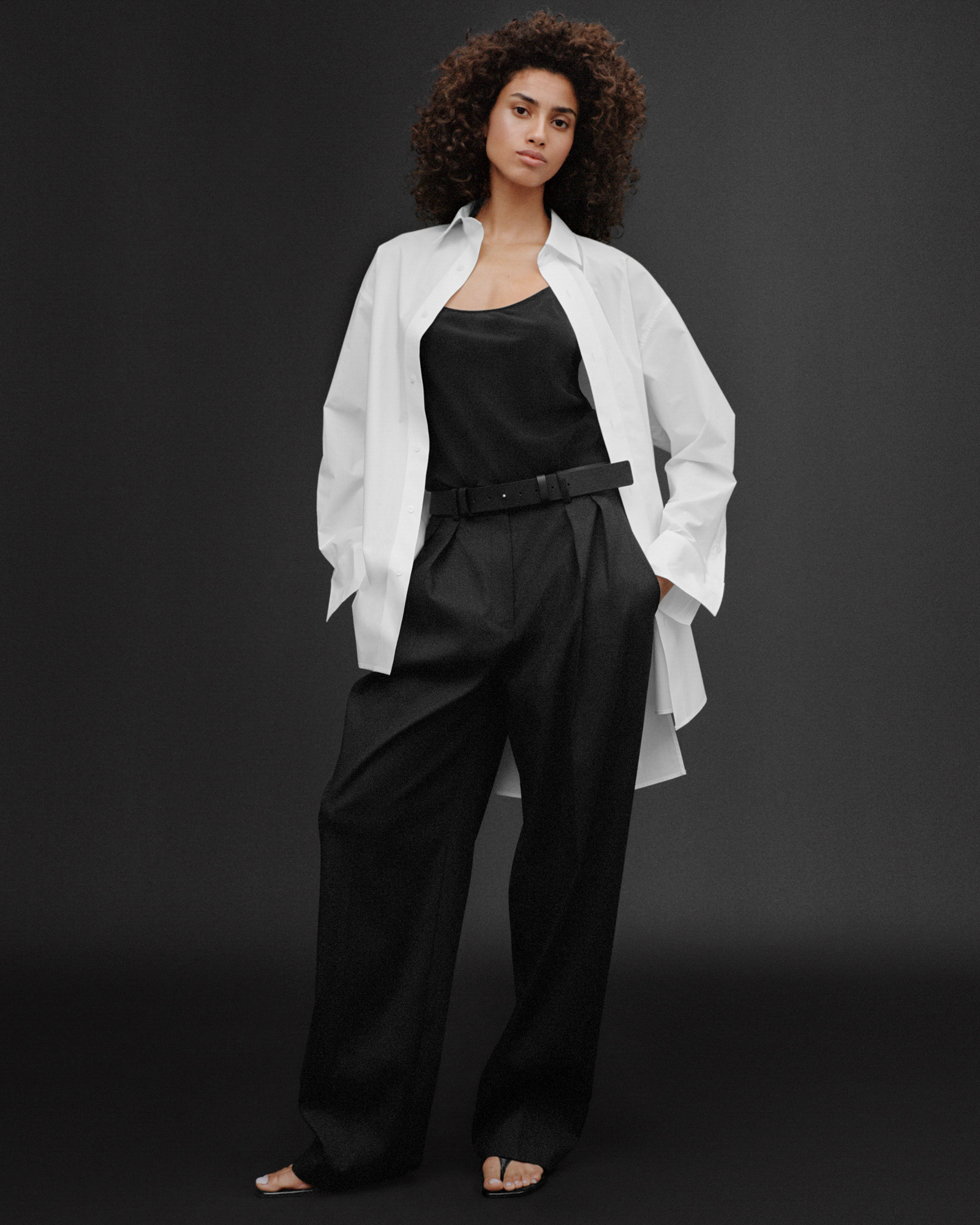 A woman is standing against a black background. She is wearing a black top, and black trousers. Overtop she has on a white button up shirt. 