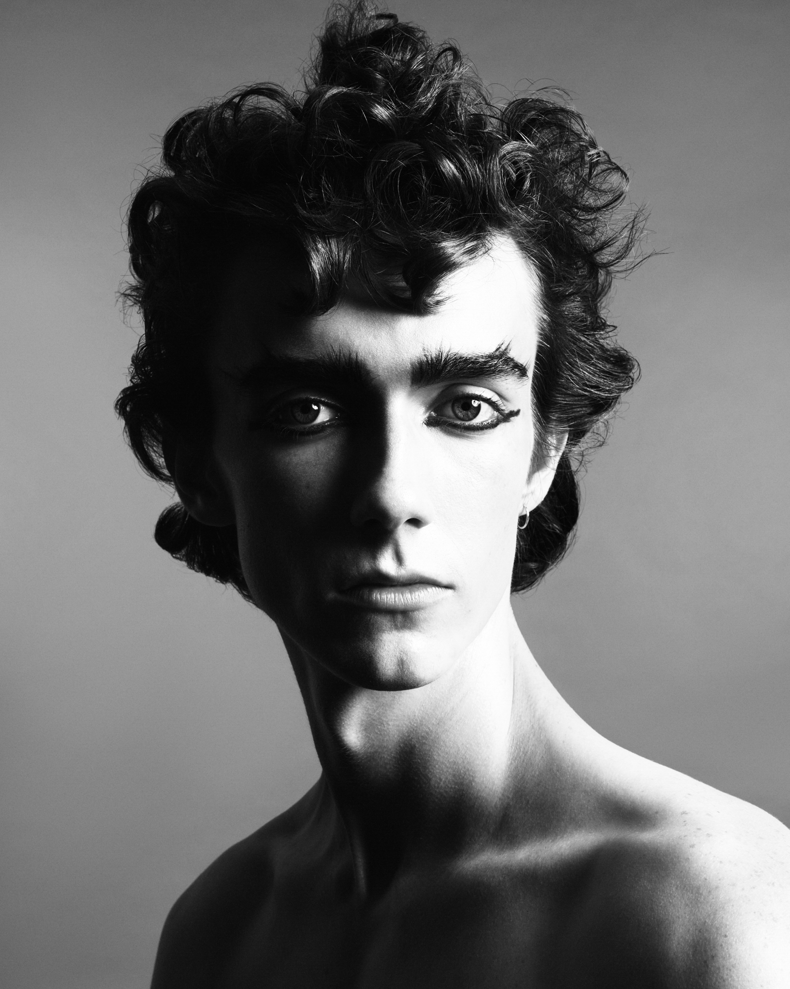 A male model is in the center of the frame, the image has a lot of contrast where only half of the model's face is in the light. They have thick black eyeliner, and thick curly hair that is styled up. Bare shoulders are visible. 