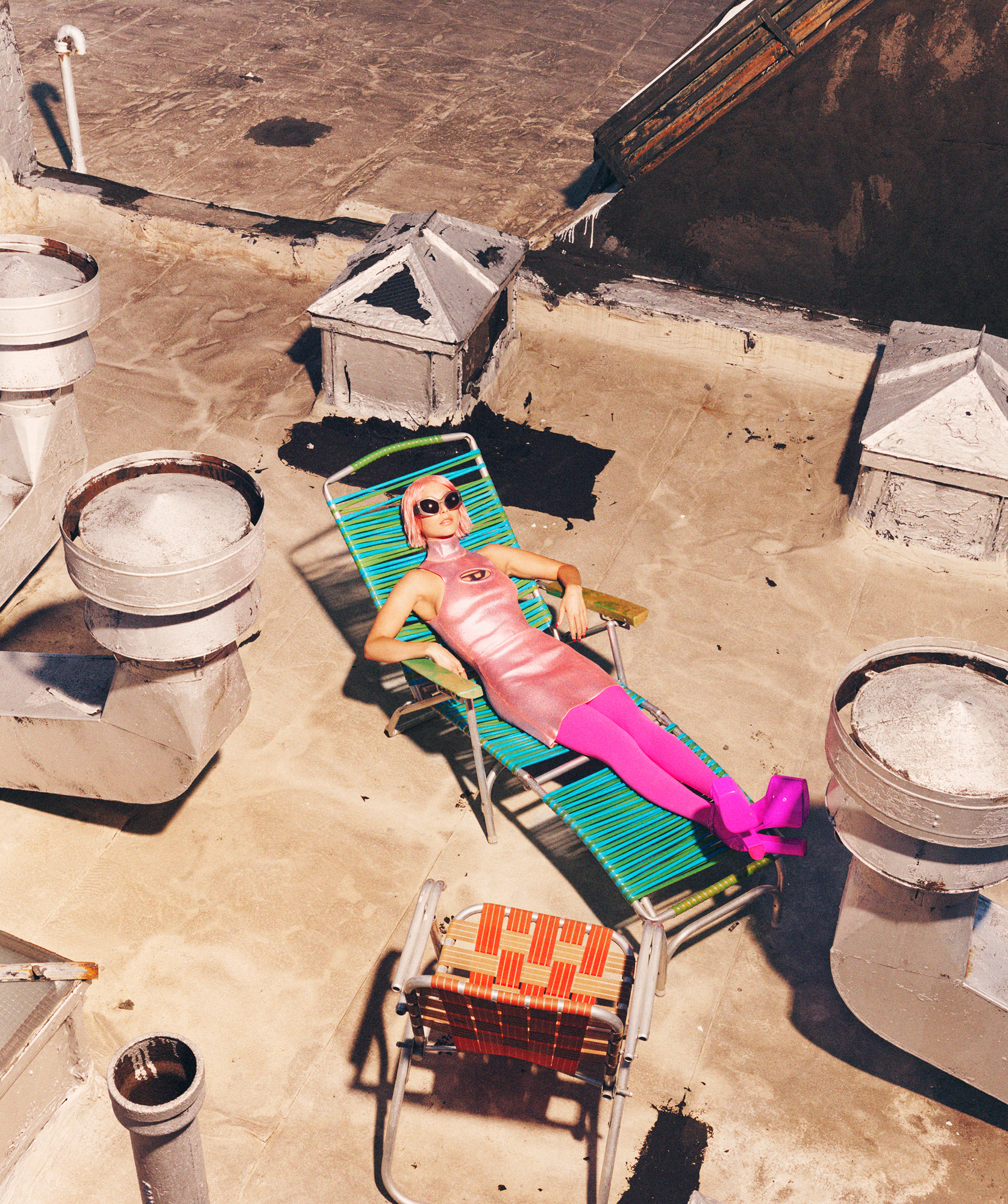 Emma Chamberlain is wearing a pink Diesel dress with neon pink leggings and shoes. She is reclined on a pool chair that is green and blue. An orange deck chair is empty in front of her. The whole scene is on a city rooftop. 