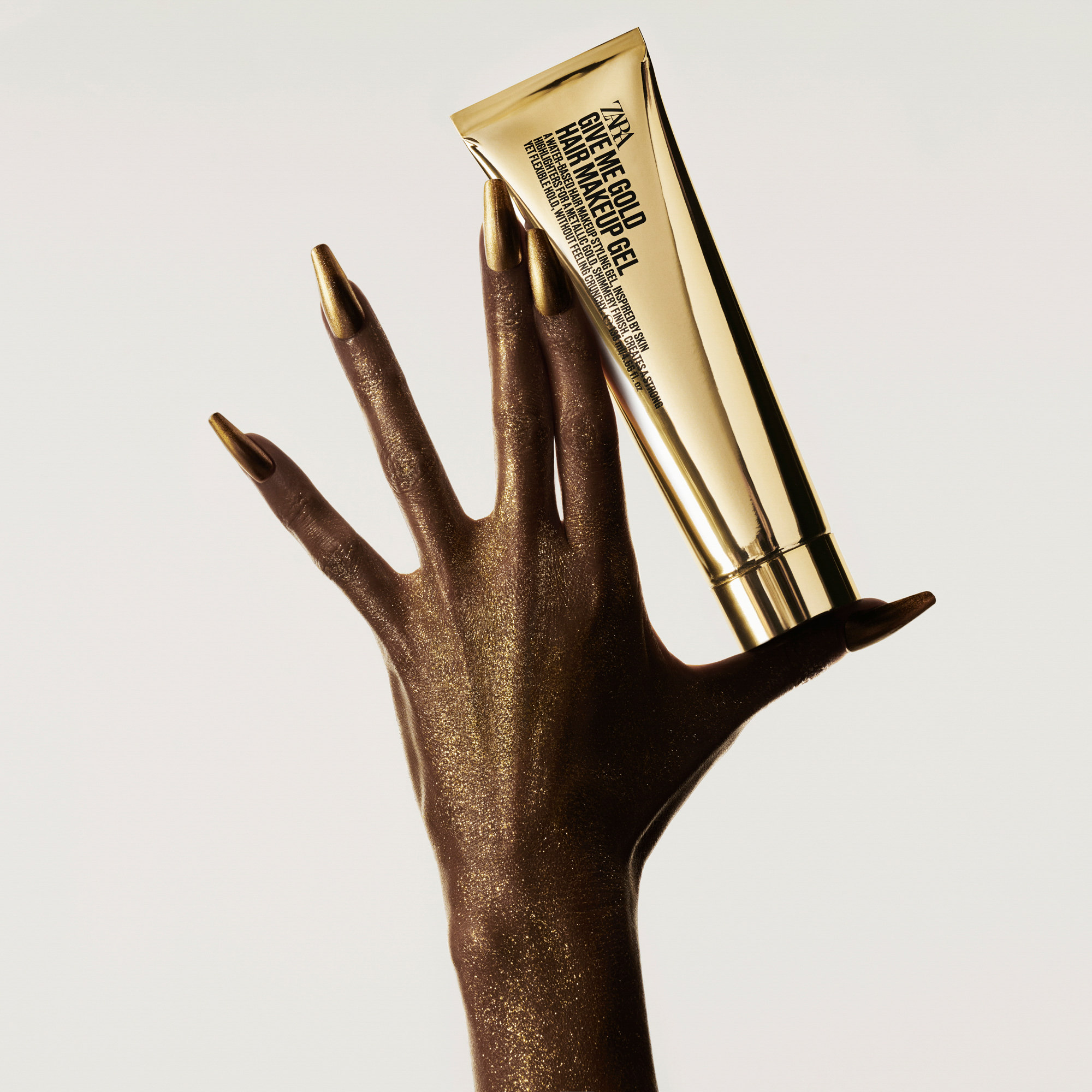 A gold paint covered hand with golden nails has the fingers spread out holding up a Zara hair makeup gel product. 