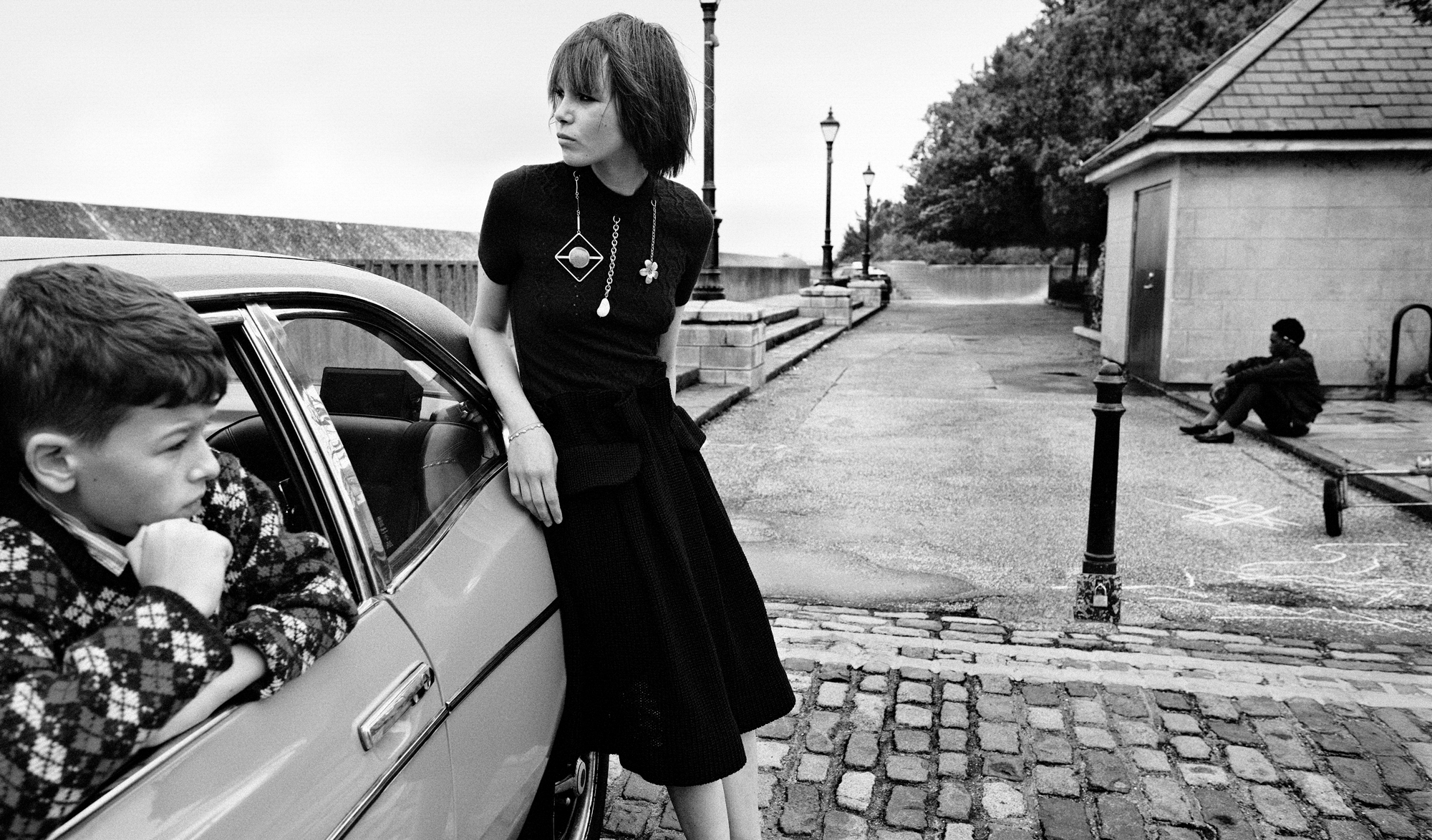 A woman wearing a black dress is leaning against a car that is parked on a cobble stone road. She is looking over her shoulder in the distance. A young boy is leaning out one of the car windows, and in the distance another boy is seated on a street curb. 