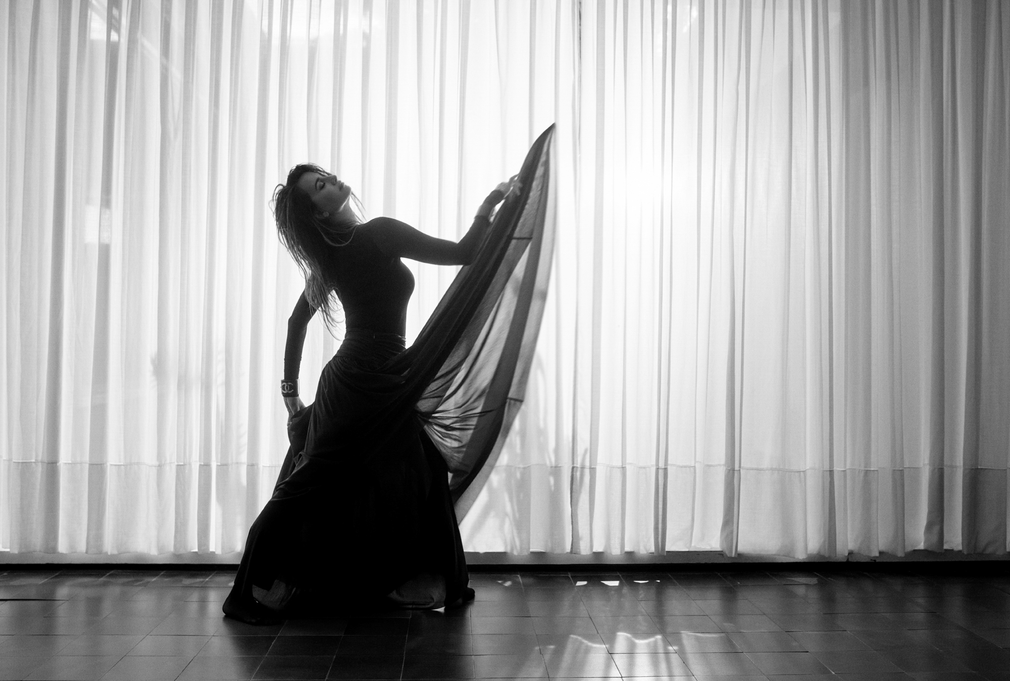 Penelope Cruz is dancing in a full length dress within an interior room with tiled floors and sheer curtains. She is leaning her head back while throwing up in the air a section of her dress. 