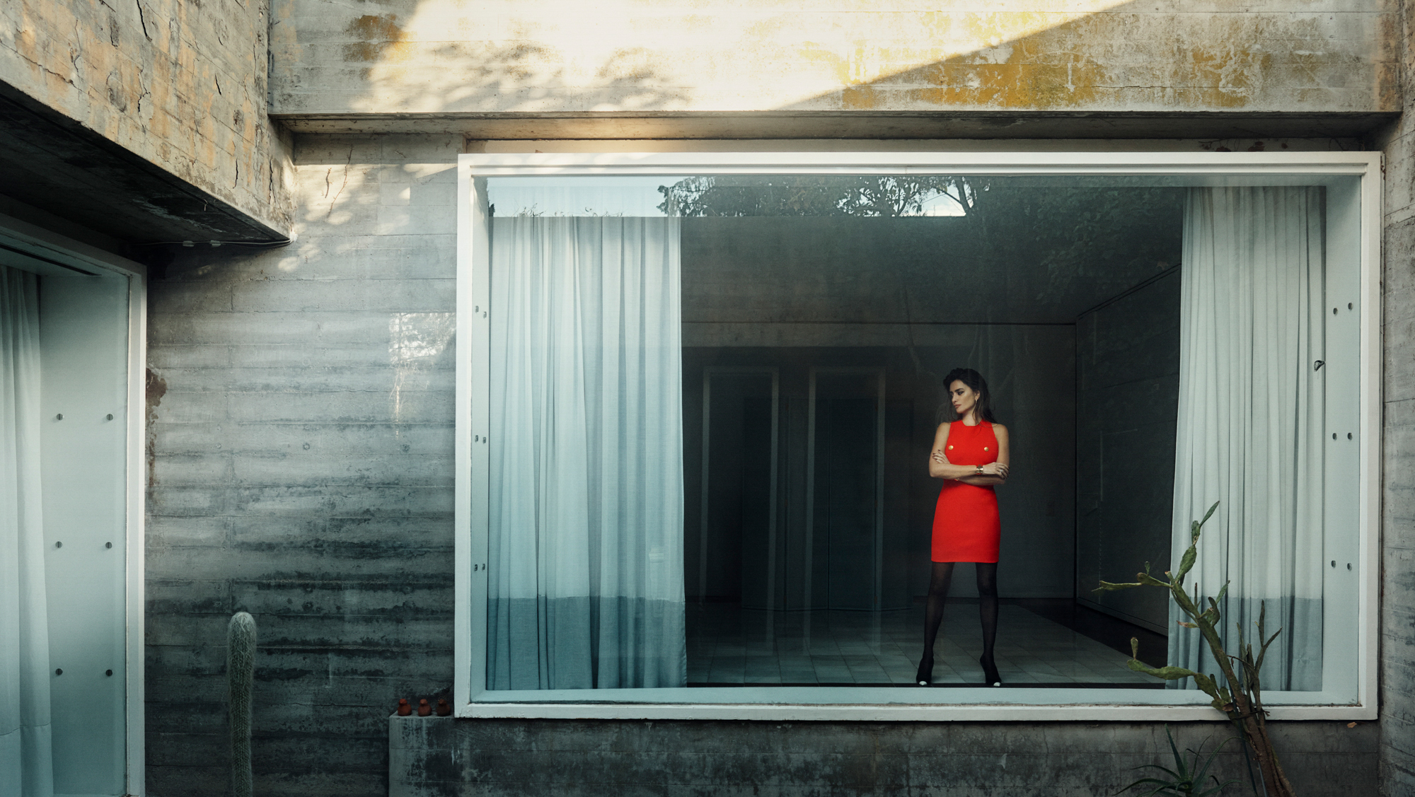 Penelope Cruz is seen standing within a large window wearing a vivid red dress and black stockings. She has her arms crossed and is looking slightly down towards the left of the frame. 