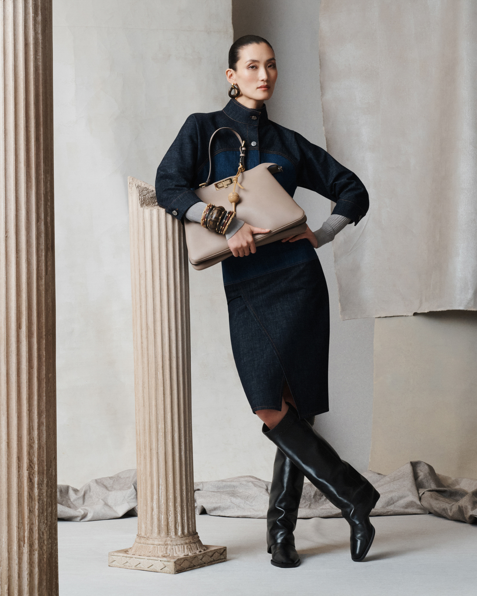 A woman is standing in a knee length denim dress with one leg crossed over the other. She has knee high black boots, and is holding a medium sized light brown Fendi bag. Her hair is pulled back in a tight bun, and she has an assortment of brown toned accessories. She is leaning one arm up against a Greek column. The set features Greek columns, different textures of gray paper, and a gray fabric that is crumpled in a line on the floor. 