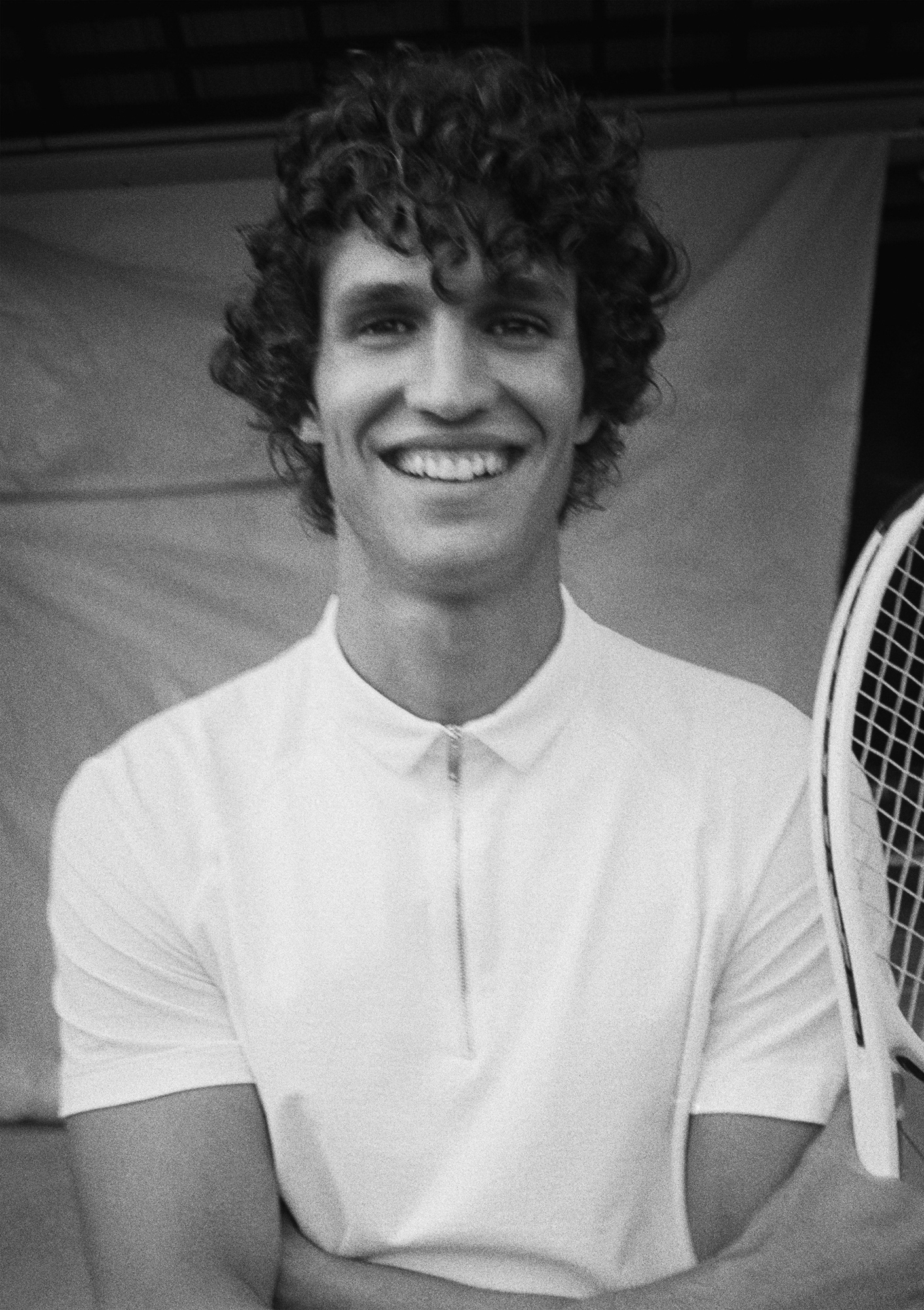A man with dark curly hair is smiling at the camera with his arms crossed. He is holding a tennis racket that is slightly visible to the right side of the frame. He is a wearing a white short sleeve shirt with a zipper, and in the background there is a fabric backdrop. 