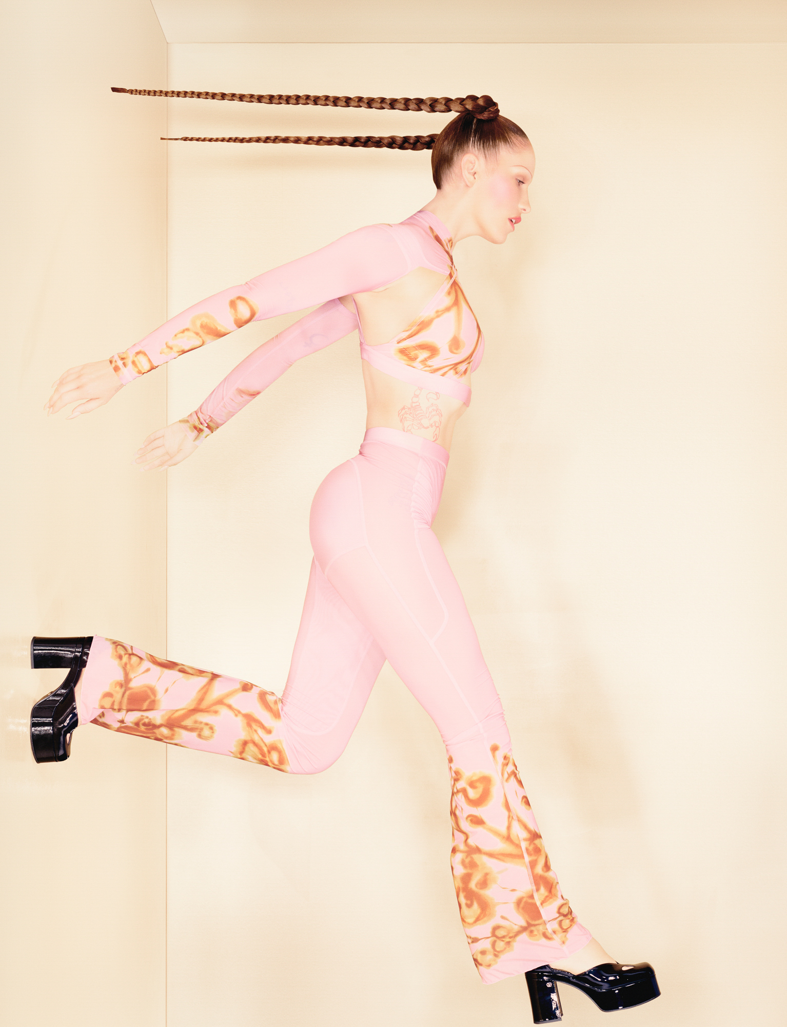 Eartheater is seen in a pink halter top with pants. The bottoms of the sleeves and pants have orange swirl patterns. She is wearing patent leather heels, and her hair is in tight long pigtail style braids. It looks at if she is leaping off the left hand side of the wall. The room has plain walls and a light orange coloring. 