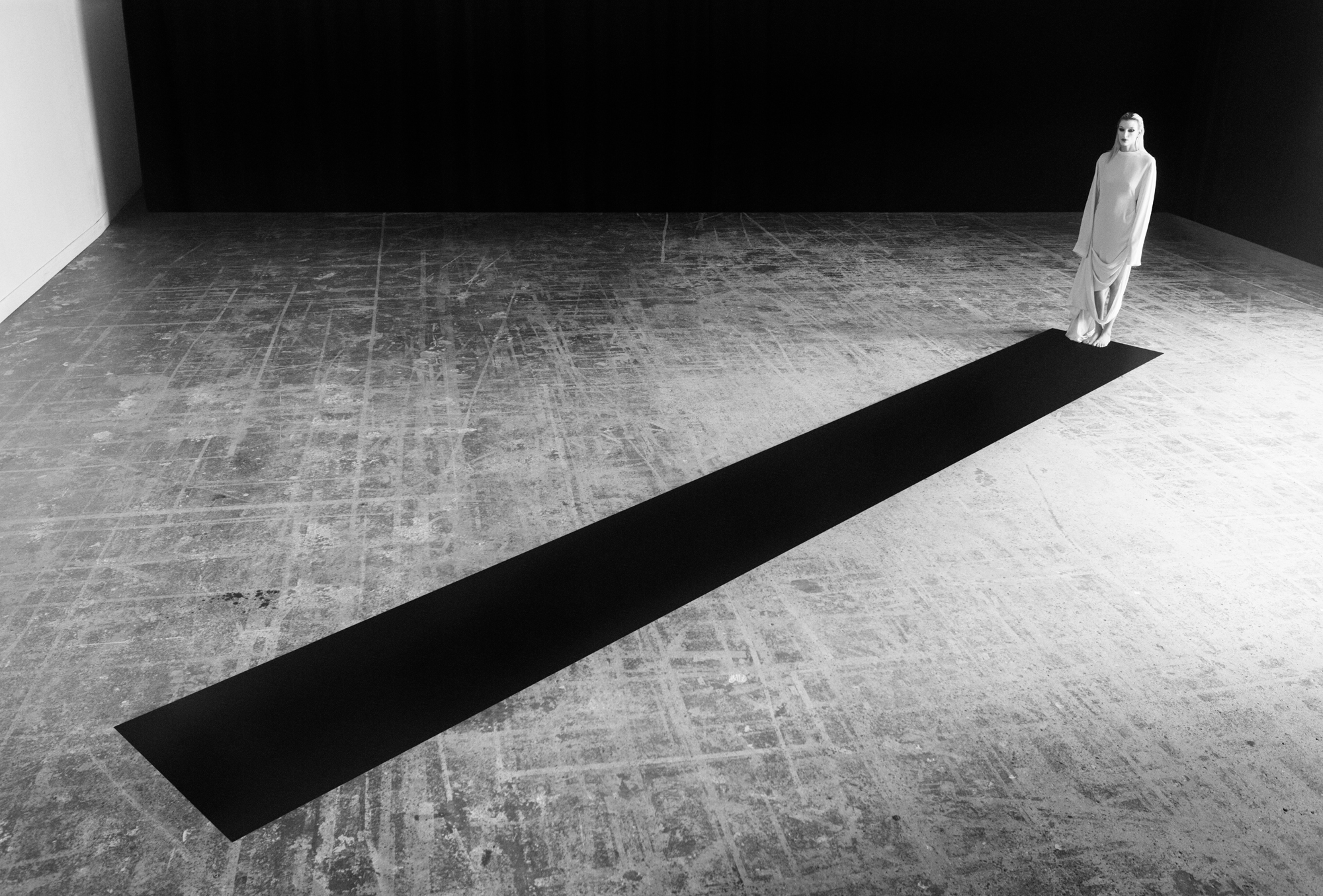 A figure dressed in white is a the top right of the frame. The figure is standing on one end of a long rectangular carpet. The camera perspective is looking down on the scene. 