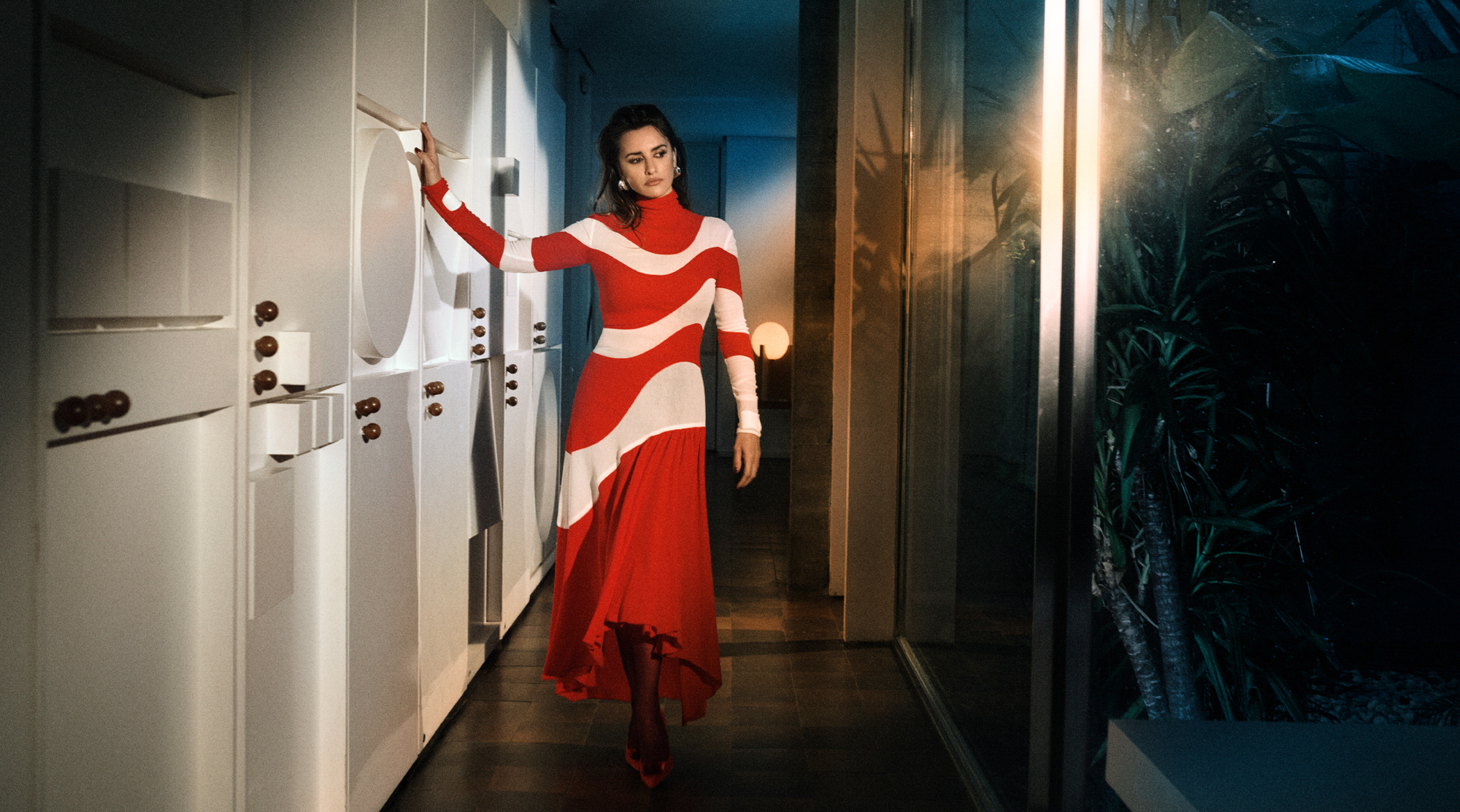 Penelope Cruz is walking through an interior hallway that has floor to ceiling windows on the right side of the frame. She has one hand extended to the wall next to her. She is wearing a red dress with white swirled lines that are apart of the design of the dress. 