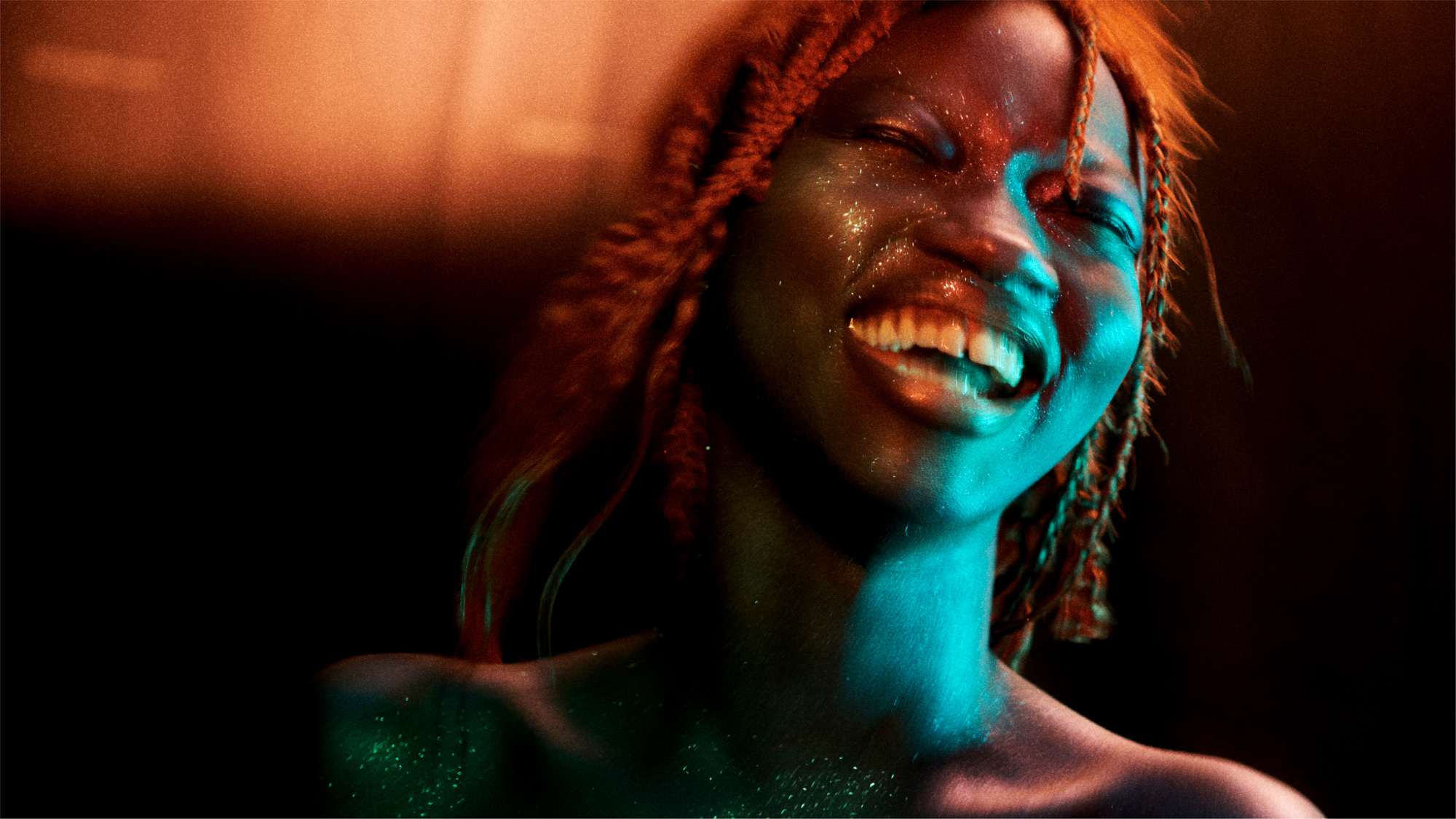 A woman has her head back laughing, light blue light is shining on her face from the right side of the frame. She has braids, glitter on the face, and shoulders exposed. Warm light is coming in from the top of the frame. 
